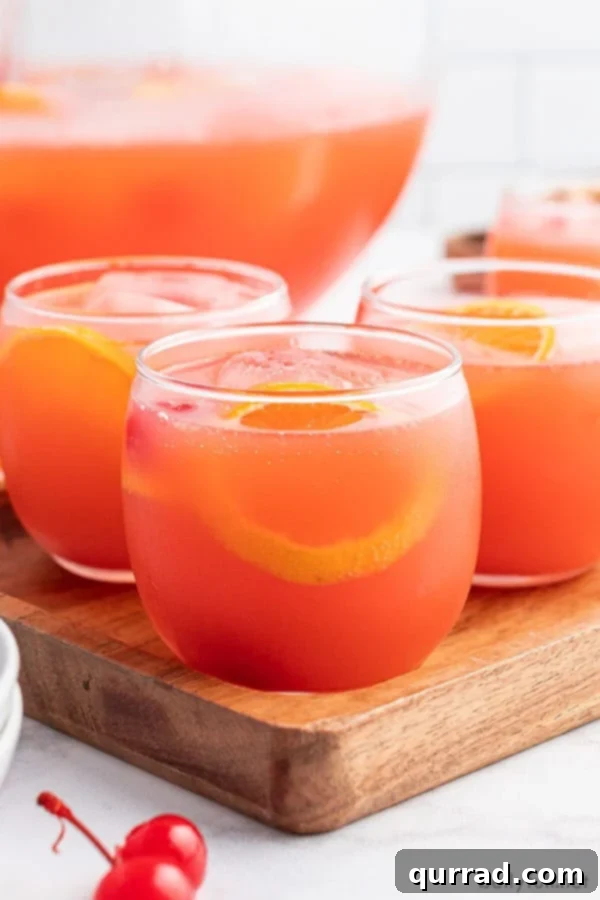 A close-up of Shirley Temple Party Punch served in a clear glass tumbler, garnished with a maraschino cherry and an orange slice.