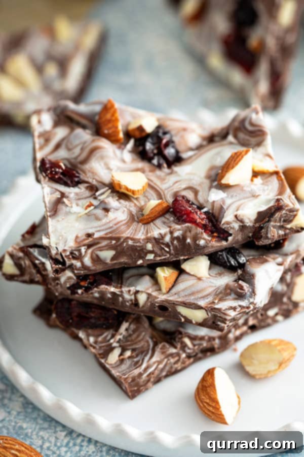 Sweet-Tart Cranberry Almond Chocolate Bark 2 Three pieces of almond chocolate bark stacked on a white plate, showcasing the vibrant cranberries and crunchy almonds within the rich chocolate.