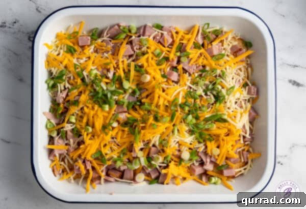 Layers of shredded cheese, diced ham, and fresh scallions evenly distributed over the hash brown patties in a casserole dish, prior to baking.