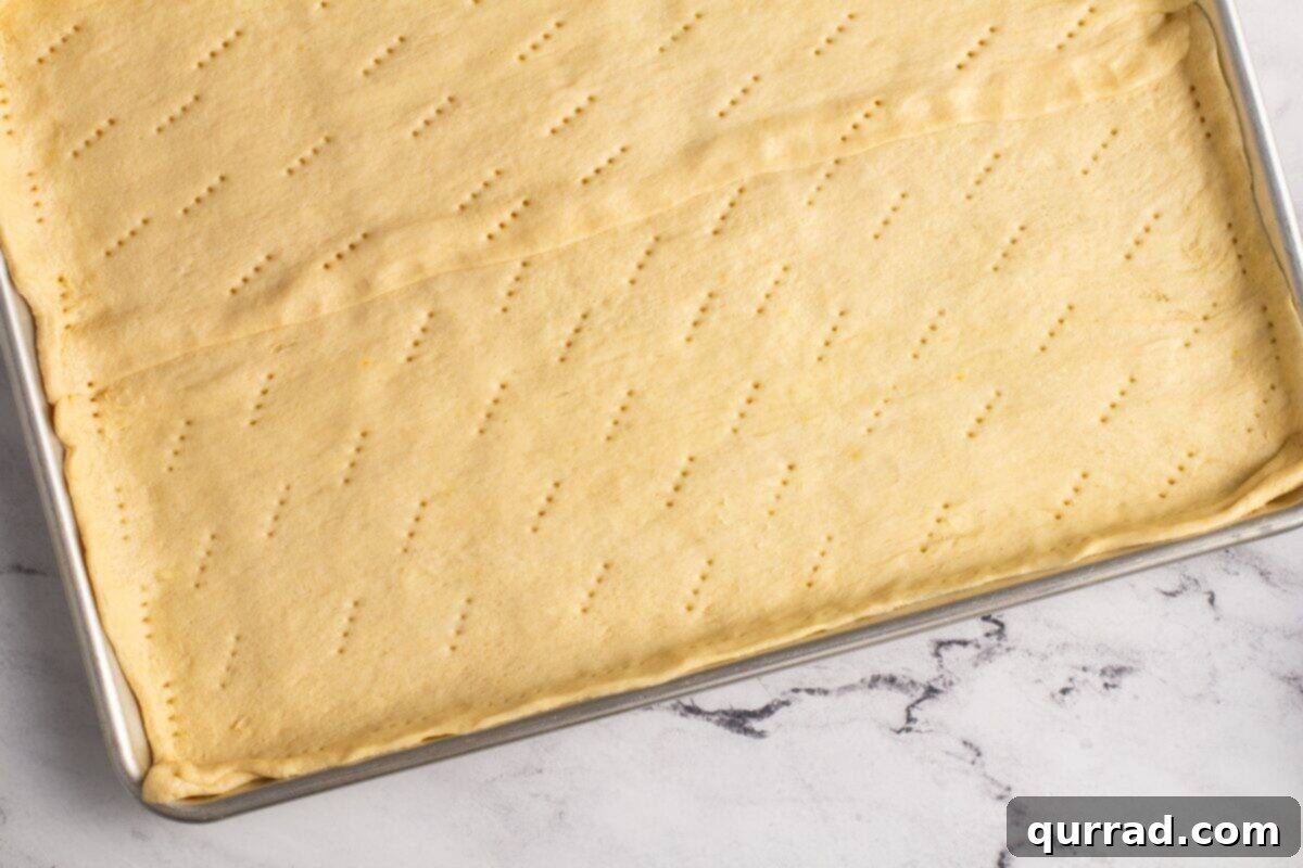 A close-up shot of crescent roll dough unrolled on a baking sheet. The dough is stretched to cover the pan, seams are pinched together, and small, evenly spaced holes have been poked across its entire surface with a fork, ready for baking.