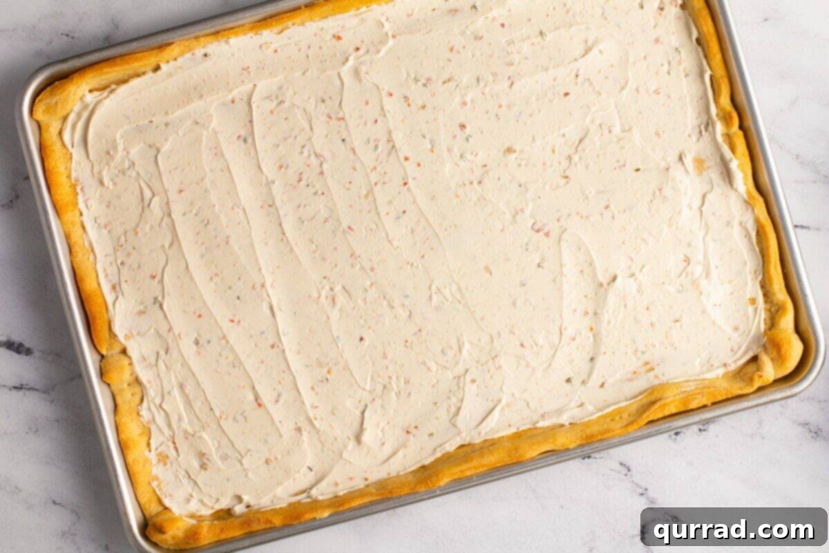 A perfectly baked and cooled crescent roll crust sits on a baking sheet, now generously covered with a smooth, even layer of garden veggie cream cheese spread, ready for the next layer of toppings.