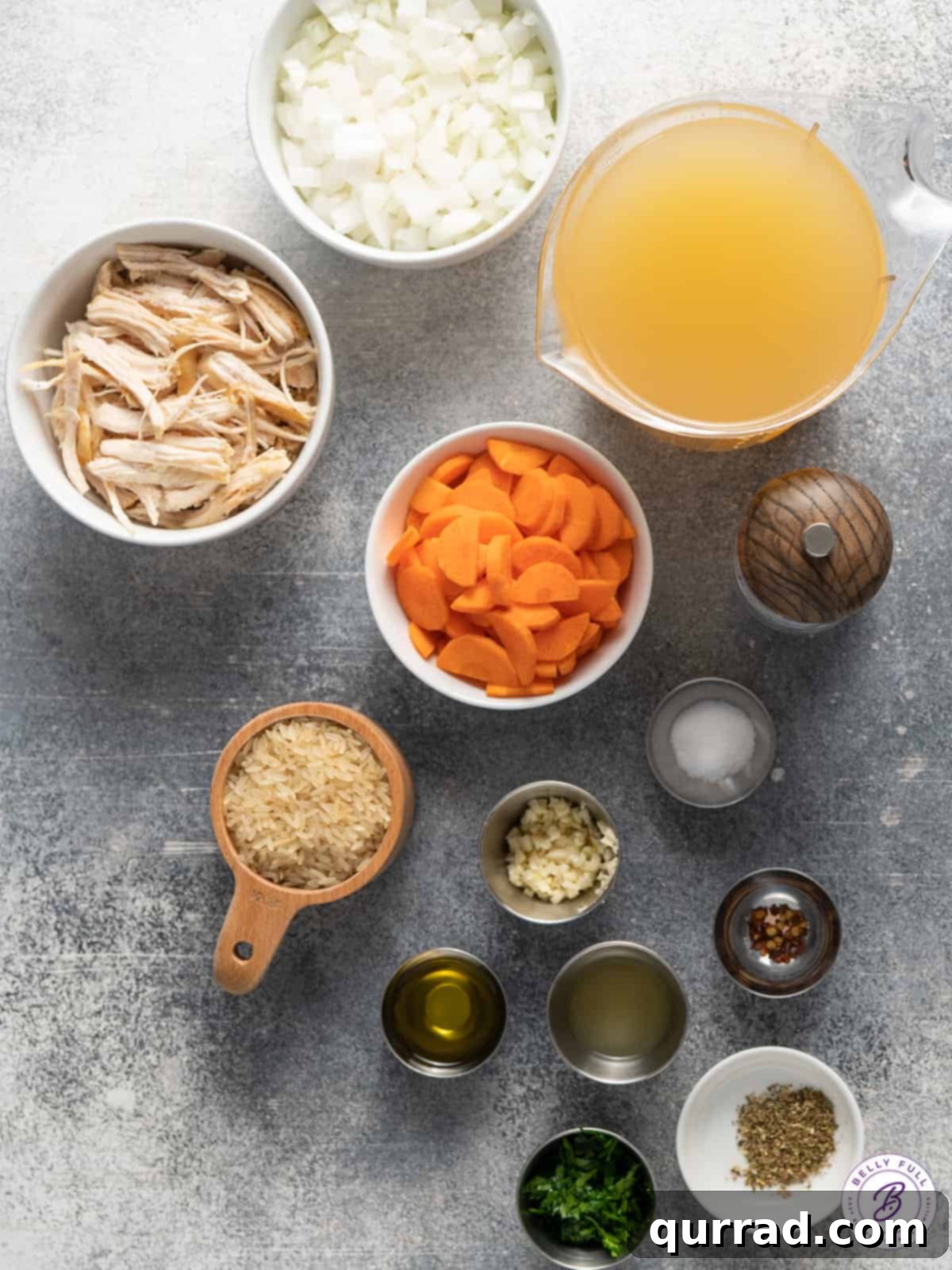 Golden Chicken Rice Elixir 3 A collection of fresh ingredients laid out for chicken and rice soup, including diced onions, sliced carrots, garlic cloves, a carton of chicken broth, and uncooked long-grain white rice.