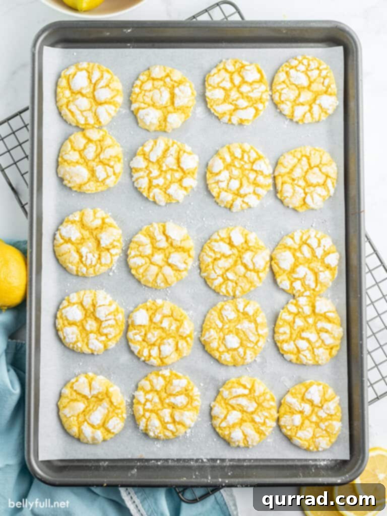 Freshly baked Lemon Crinkle Cookies cooling on a baking sheet lined with parchment paper, their powdered sugar coating creating delicate cracks and a vibrant yellow hue.