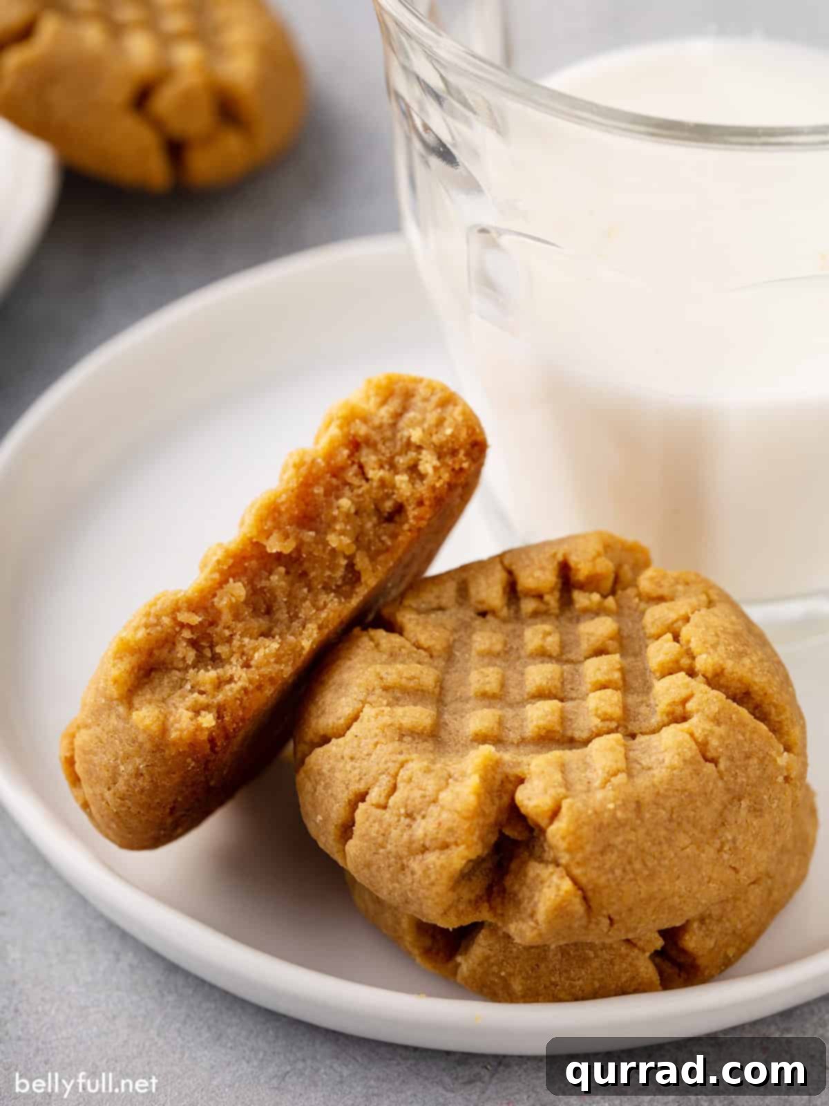Three perfectly baked peanut butter cookies arranged on a white plate, two whole and one split in half, showcasing their chewy interior.