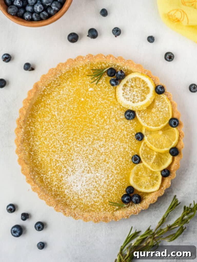 A beautifully presented whole lemon tart, garnished with delicate lemon slices, fresh blueberries, and a dusting of powdered sugar, perfect for any occasion.