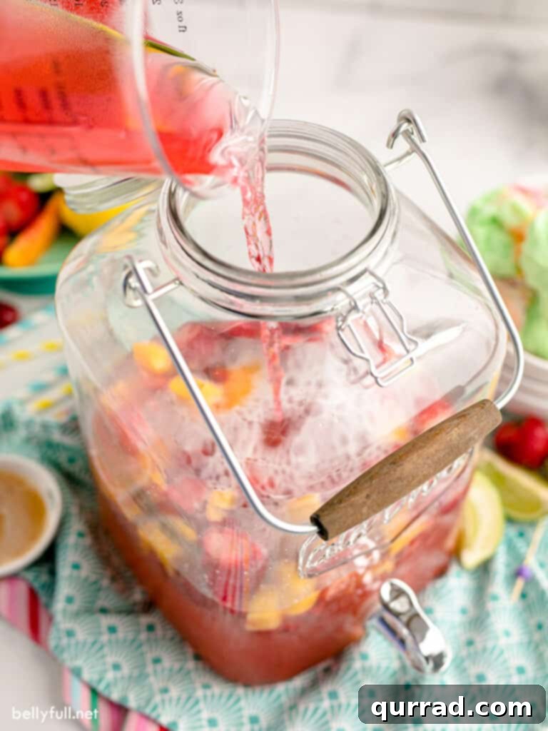 A large clear punch dispenser being filled with a colorful fruit punch mixture, showing frozen fruits settling at the bottom.