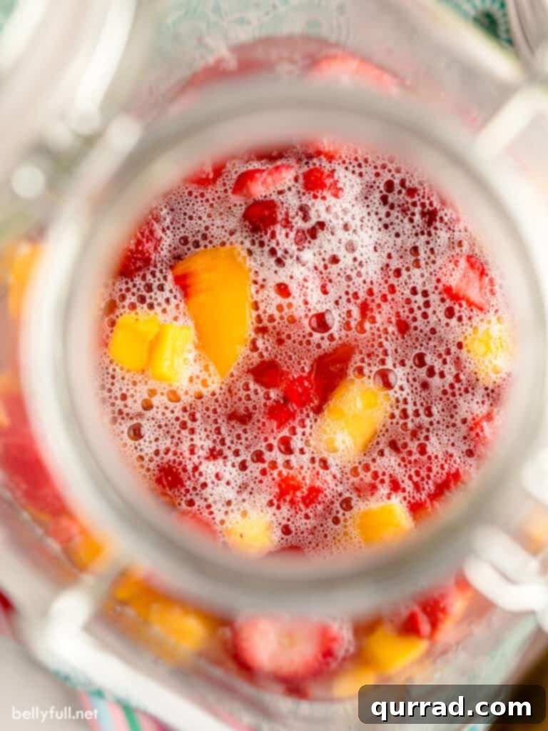 An overhead view of a large punch dispenser filled with vibrant sherbet punch and various frozen fruits, ready for a party.