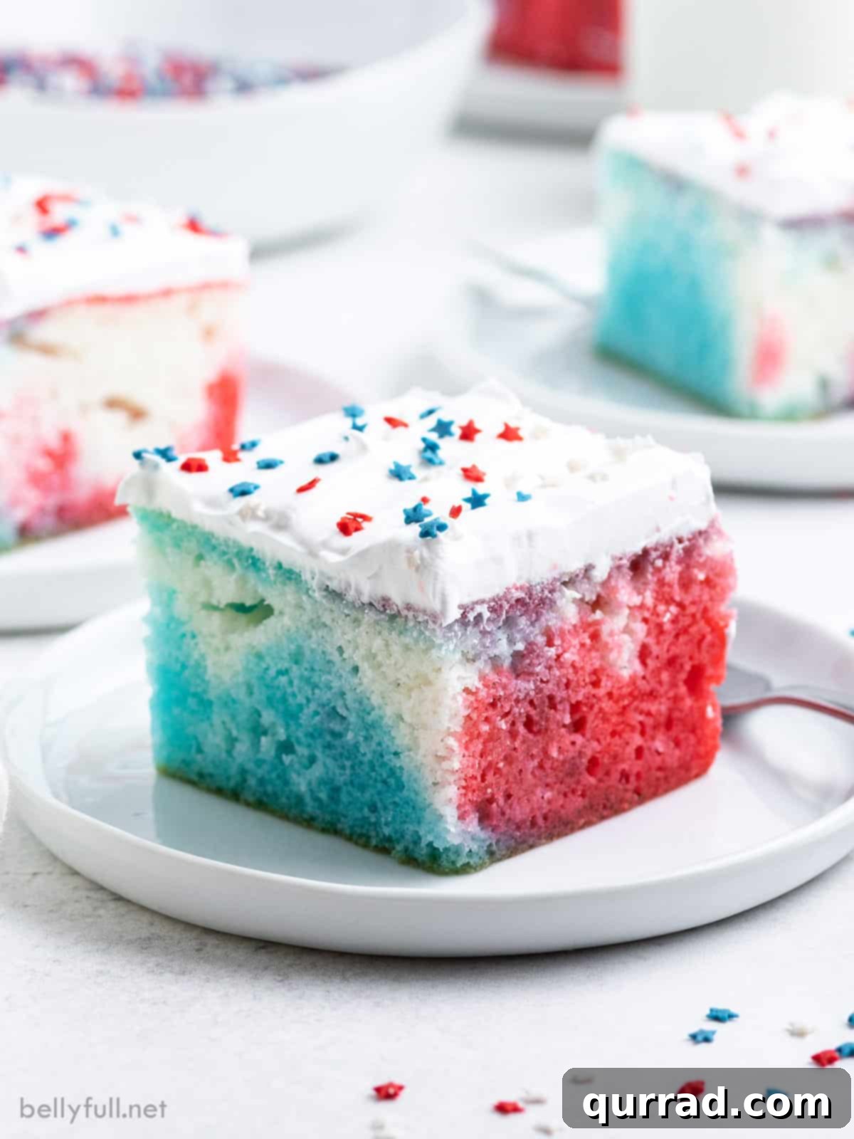 Festive Flag Poke Cake 2 A vibrant slice of Red, White, and Blue Poke Cake, showcasing distinct layers of red, white, and blue, presented on a pristine white plate, garnished with patriotic sprinkles. This easy, festive, and moist Jell-O poke cake is ideal for 4th of July parties.