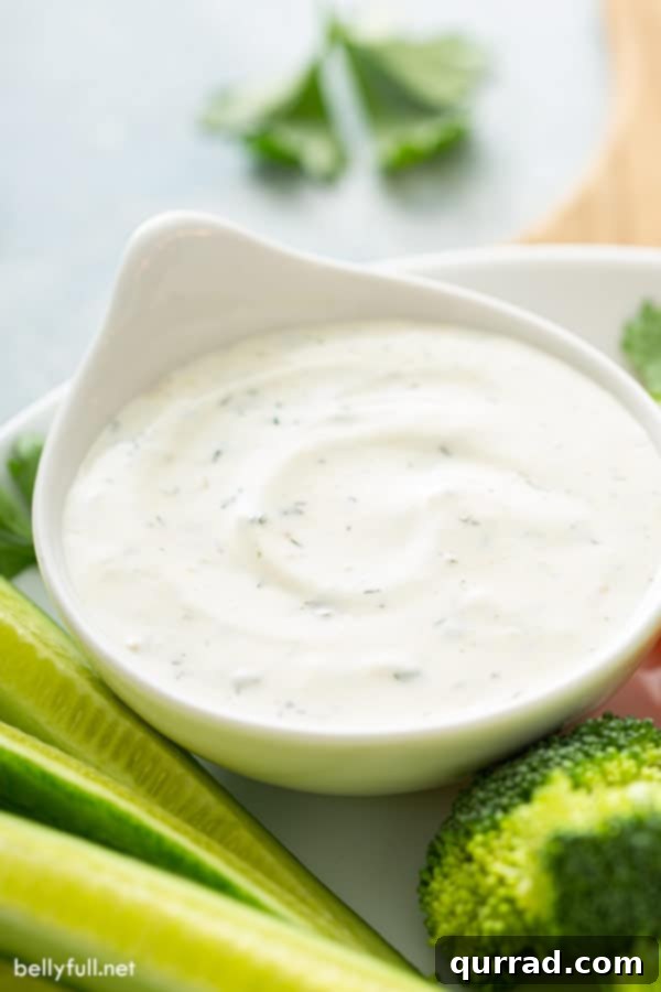 A white bowl of homemade ranch dip with cucumber spears and broccoli on a plate, showcasing its fresh appeal.
