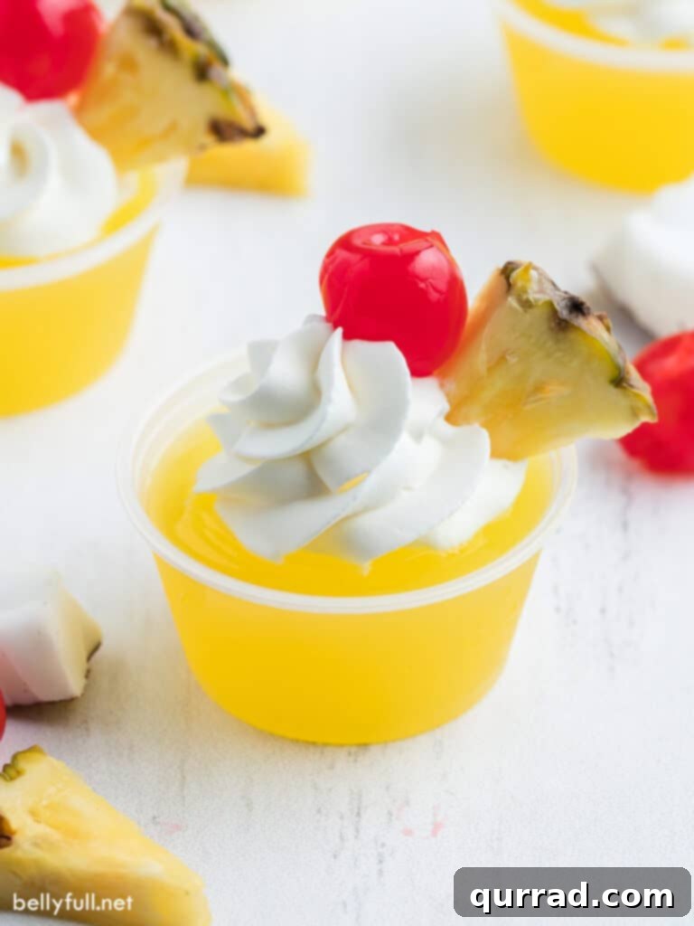 Pineapple Coconut Rum Jellies 2 Refreshing Pina Colada Jello Shots garnished with fresh whipped cream, a mini pineapple wedge, and a vibrant maraschino cherry on a white surface.