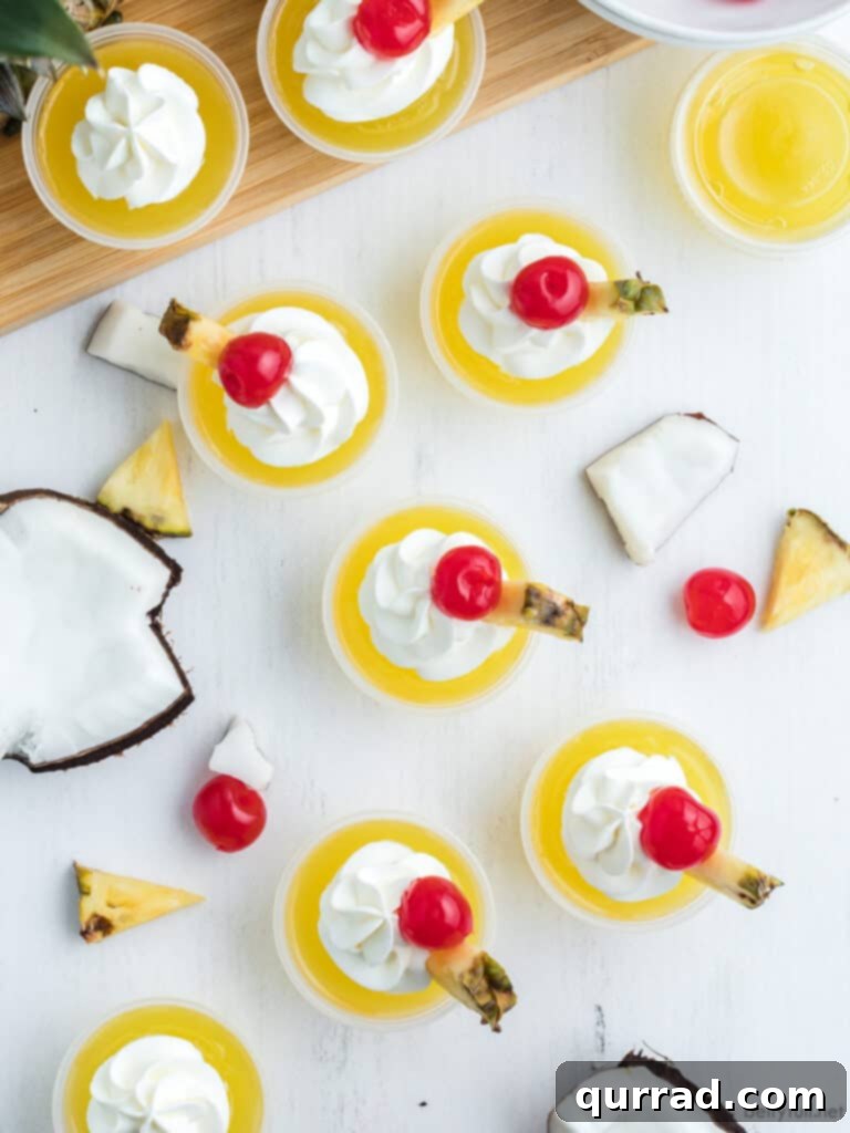 Pineapple Coconut Rum Jellies 4 Overhead view of multiple Pina Colada Jello Shots arranged on a white tray, each topped with whipped cream, a pineapple wedge, and a maraschino cherry.