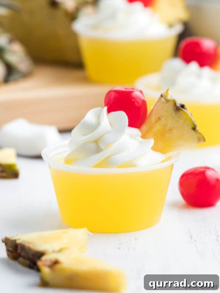 Pineapple Coconut Rum Jellies 5 Close-up of a single Pina Colada Jello Shot with whipped cream, a pineapple wedge, and a maraschino cherry on a white surface.