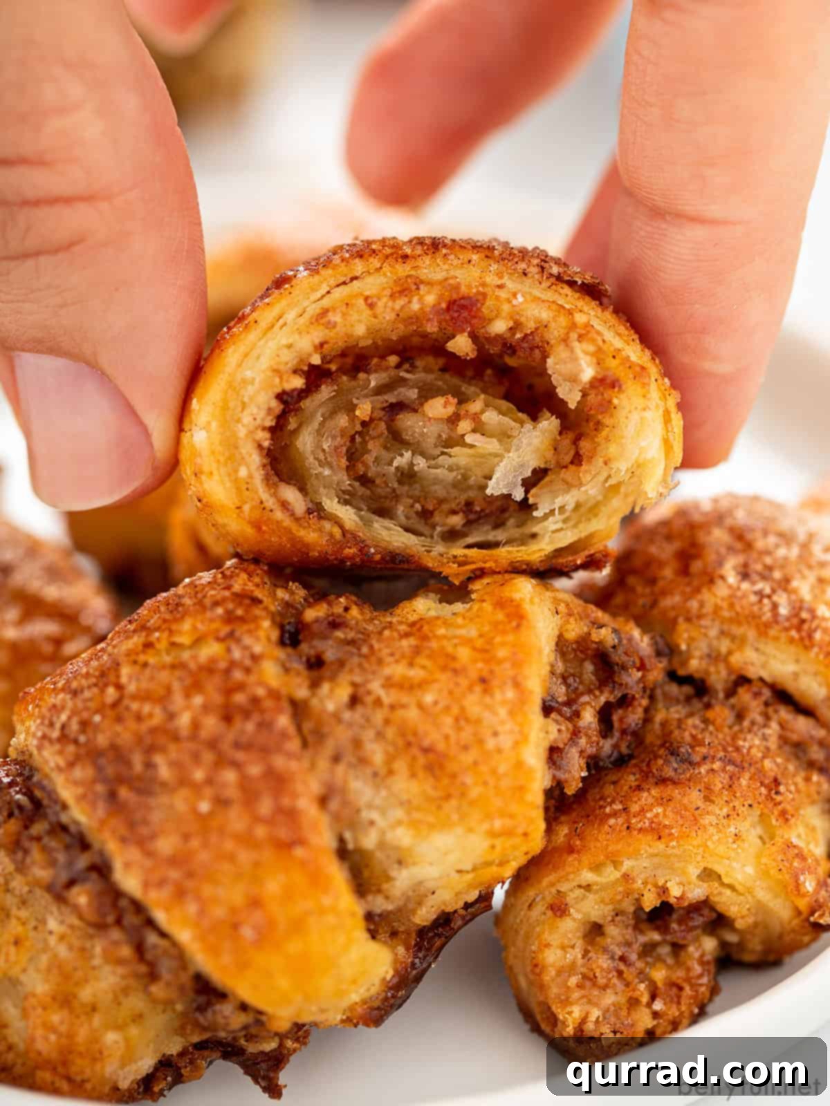 A hand reaches for a perfectly baked Rugelach cookie from a pile, highlighting its flaky layers and inviting texture.