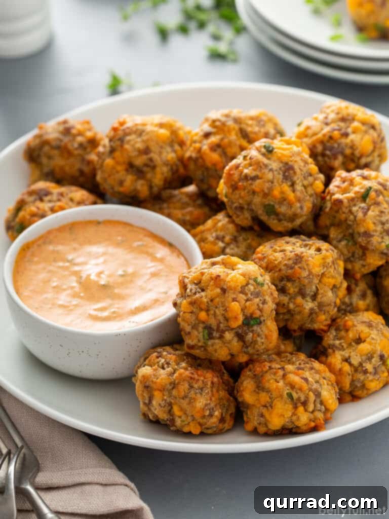 Baked, golden-brown sausage balls piled high on a white plate, served alongside a small dish of creamy remoulade sauce, ready to be enjoyed as a savory appetizer.