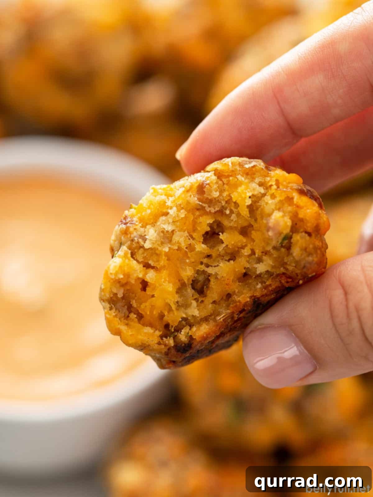 A hand holds a perfectly golden-brown sausage cheddar ball, showcasing its appetizing texture, with a blurred background of a bowl of remoulade sauce.