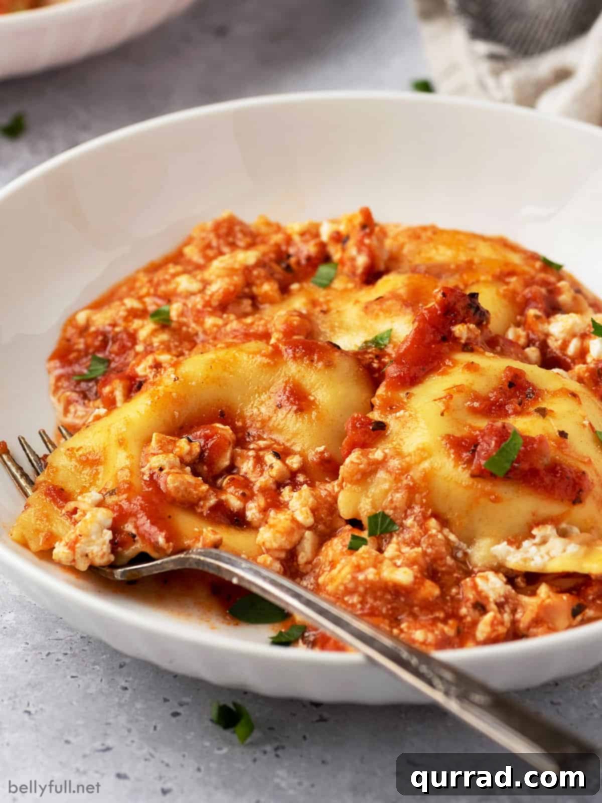 Cheesy Ravioli Dream 2 A white bowl of baked ravioli casserole with a fork on the dish, garnished with fresh parsley.