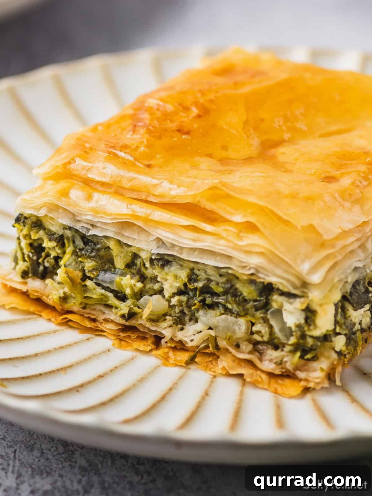 A beautifully baked slice of spanakopita on a pristine white dish, showcasing its flaky layers and vibrant green filling.
