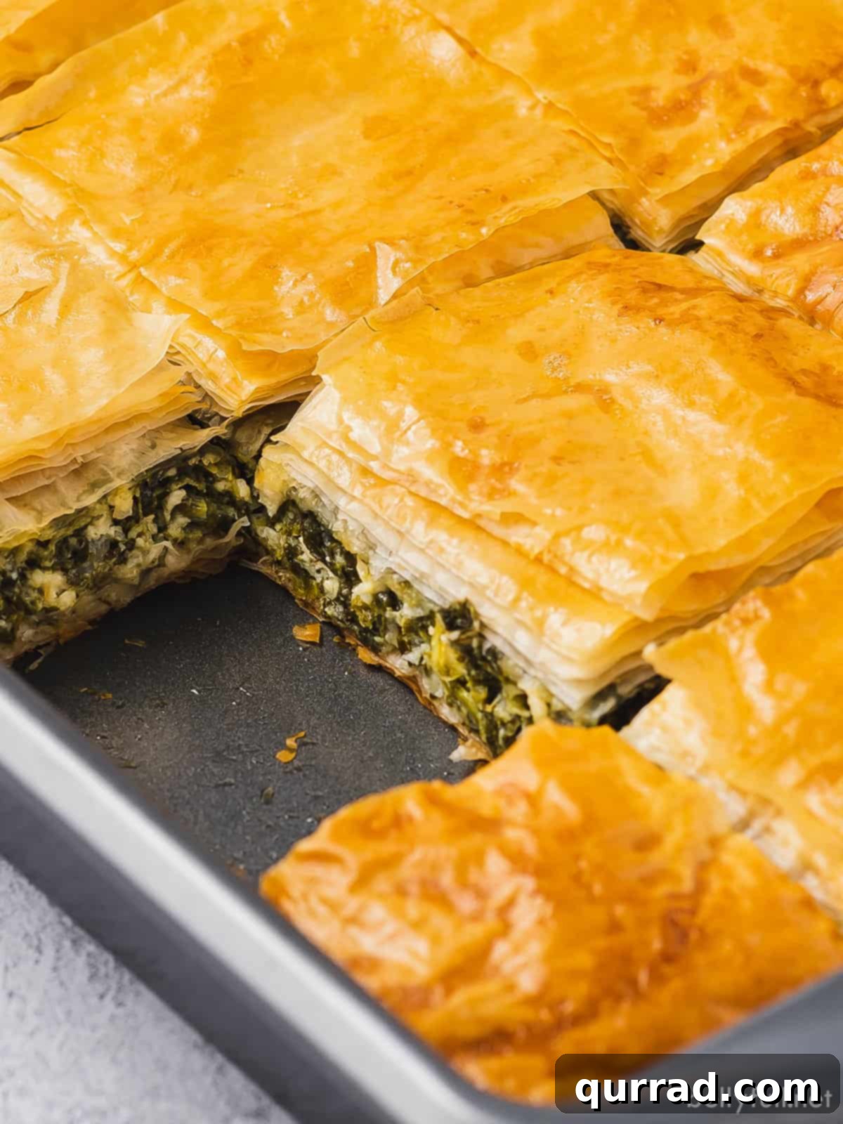 Close-up of a rustic baking dish holding a freshly baked Greek spinach pie, with one square slice expertly removed, revealing the rich filling and crisp layers.