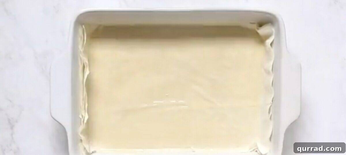 Layers of delicate phyllo dough being brushed with butter in a white baking dish, forming the base of the spanakopita.