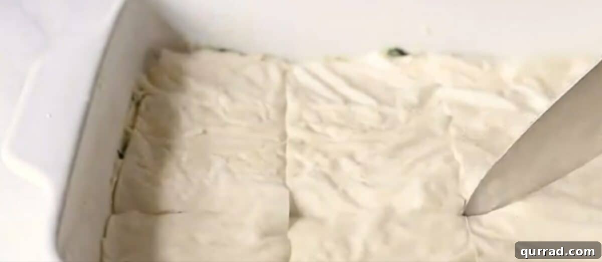 A chef's knife gently scoring the top layers of phyllo dough into squares before baking, preparing the spanakopita for easy slicing.