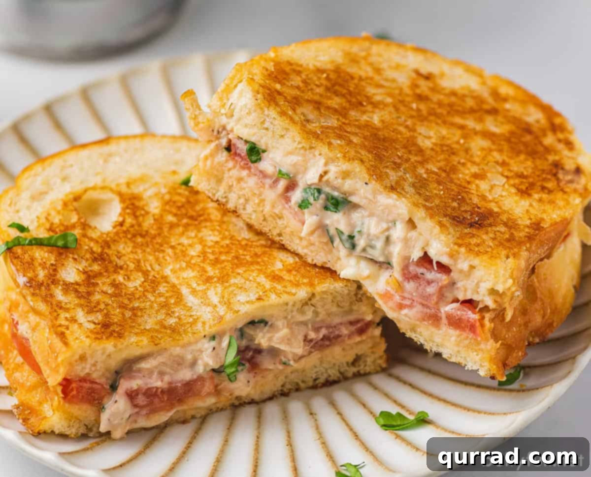 Gourmet Tuna Grilled Cheese 5 Two halves of a delicious tuna melt sandwich, perfectly grilled and revealing the melted cheese, tuna, and tomato, served on a white plate.