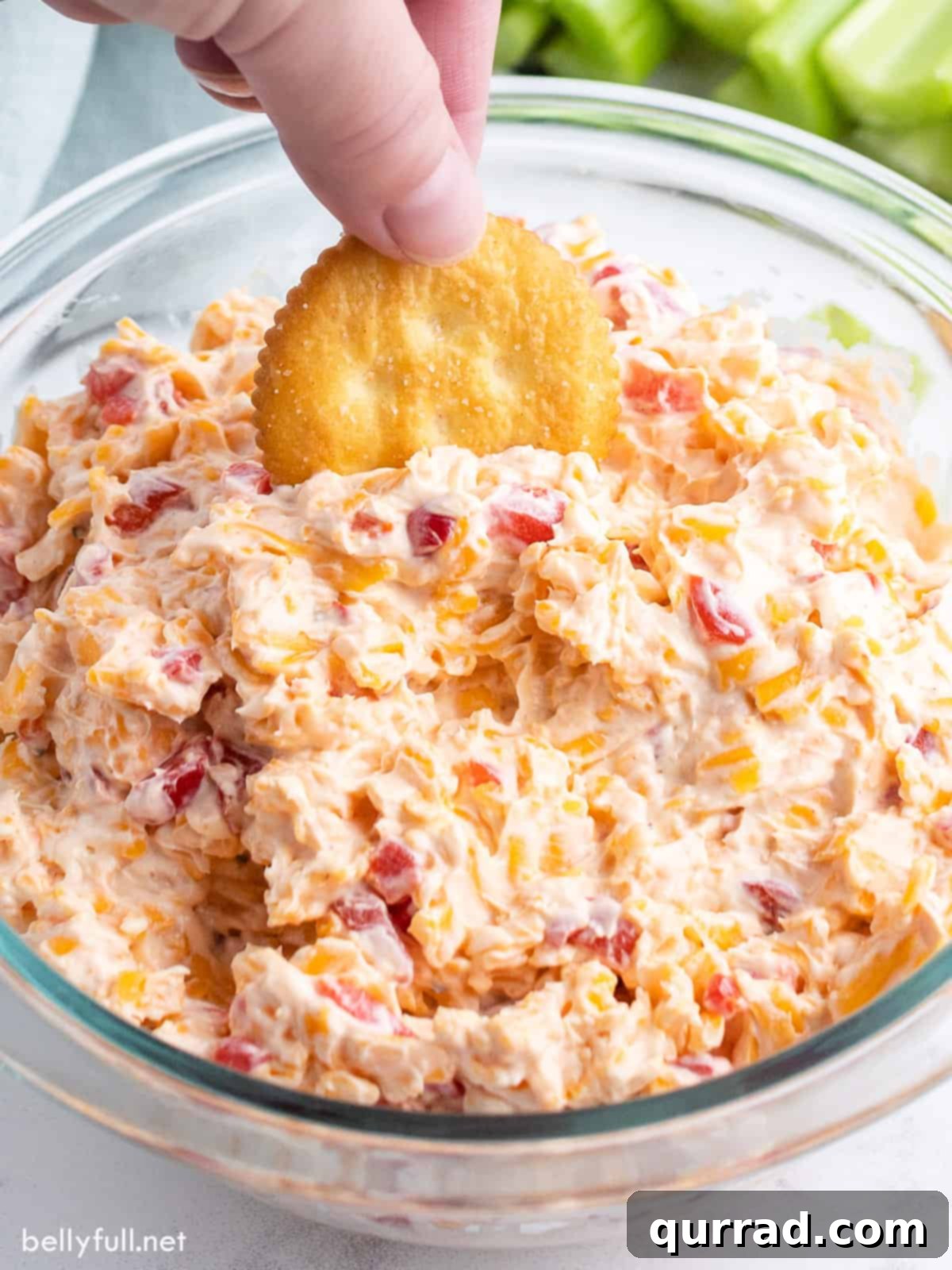 A hand dipping a buttery Ritz cracker into a bowl of creamy, vibrant pimento cheese dip, showcasing its appetizing texture and perfect consistency for scooping.
