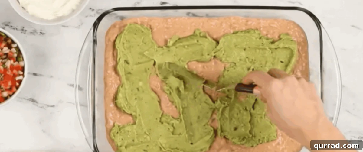 Ultimate Seven-Layer Dip 8 Spreading fresh guacamole over the refried bean layer in a dish.