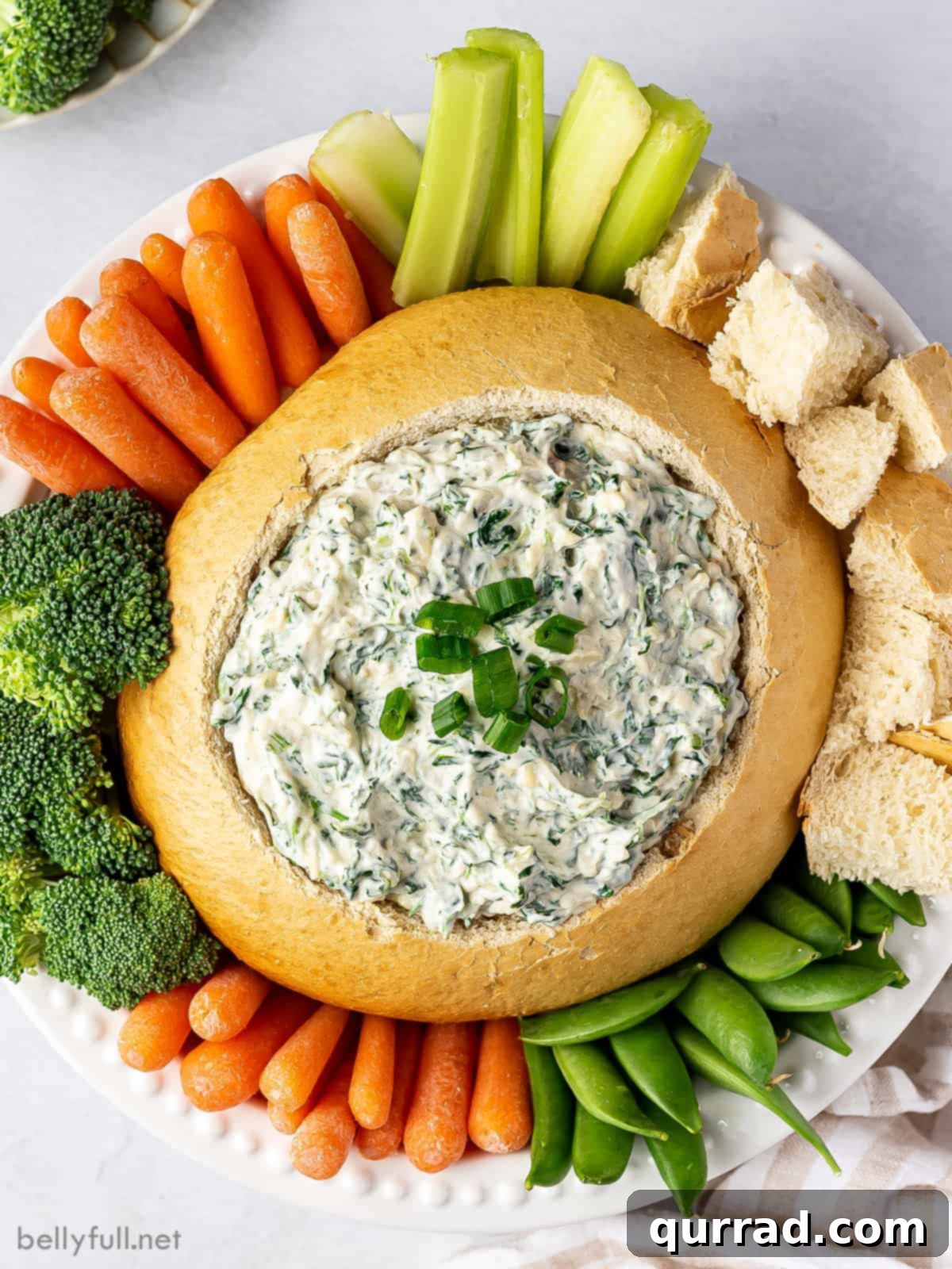 Creamy Spinach Dream 2 Overhead view of creamy spinach dip in a hollowed-out bread bowl, surrounded by fresh vegetable sticks and bread cubes for dipping. The dip has a smooth, light green texture with visible spinach flecks and is garnished with chopped scallions. Perfect for a party appetizer.