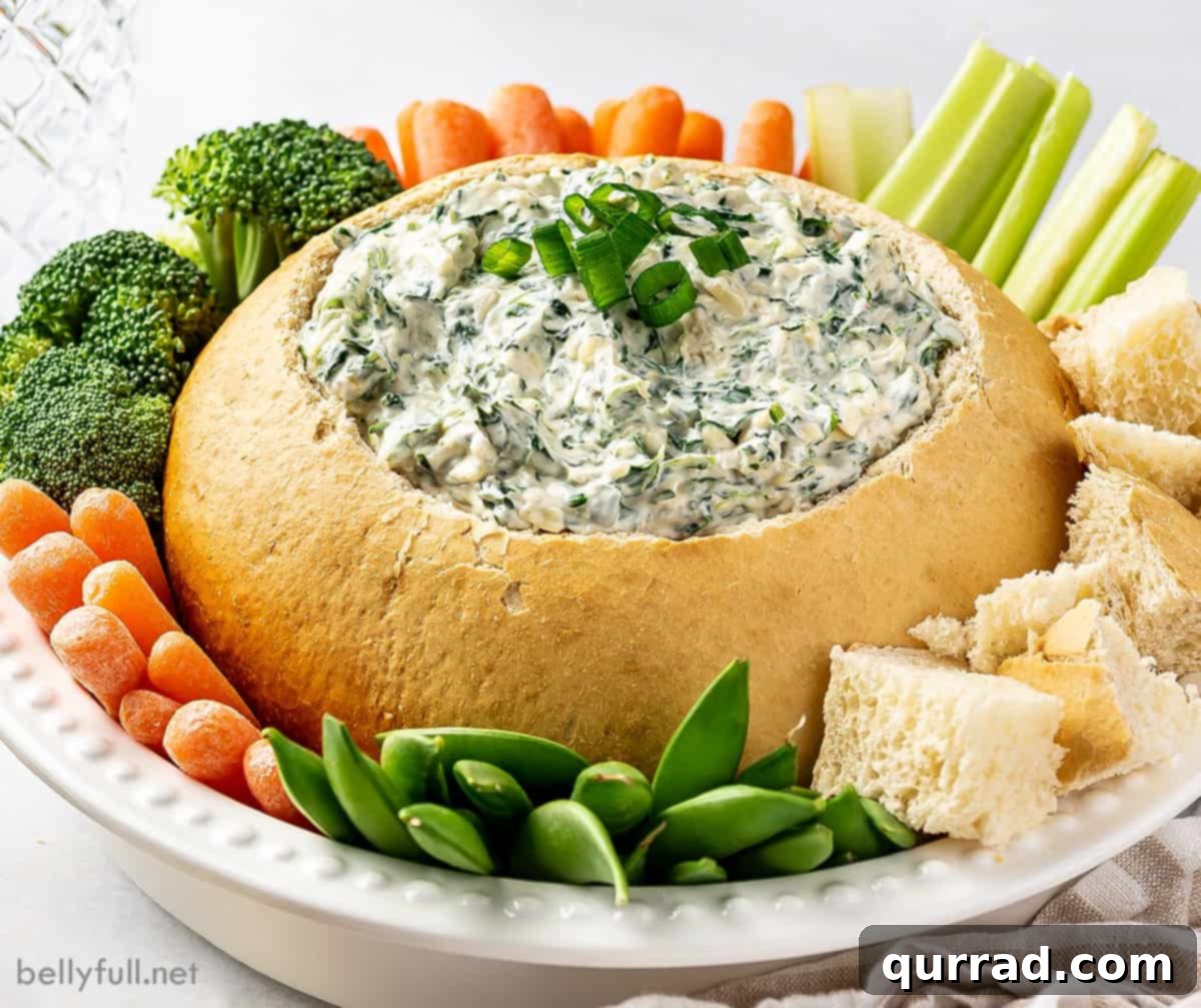 Creamy Spinach Dream 5 Angled view of a spinach dip bread bowl presented on a white plate, surrounded by an abundant spread of fresh, colorful vegetables including carrot sticks, celery, bell pepper strips, and cucumber slices. The dip looks creamy and inviting, ready for guests to enjoy.