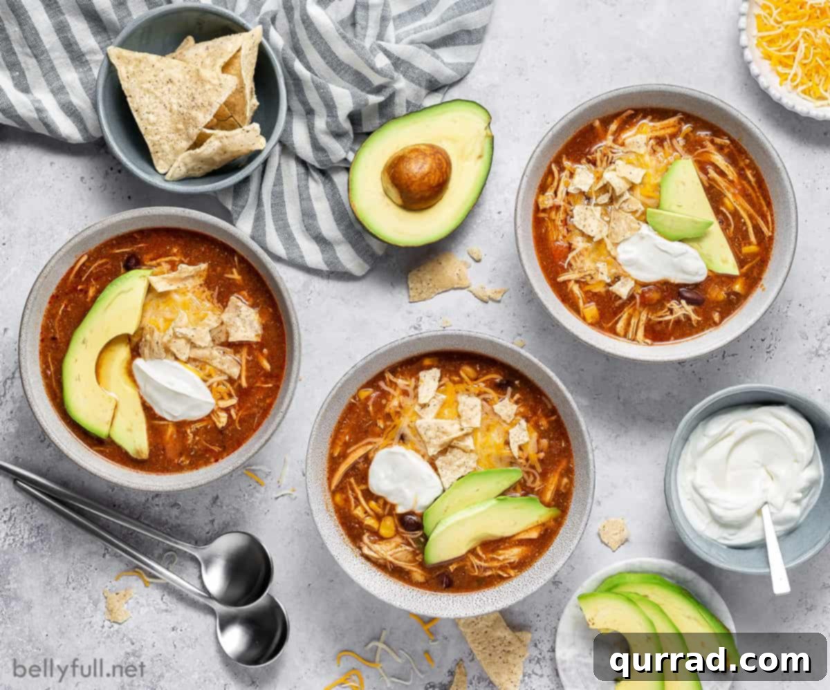 Overhead picture of 3 bowls of chicken taco soup garnished with avocado slices, half an avocado, a bowl of sour cream, a bowl of cheese, and a bowl of tortillas. 