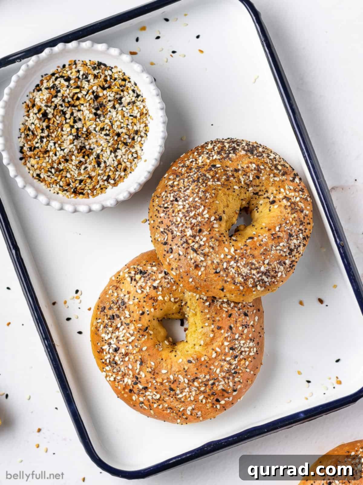 Two freshly baked bagels on a serving tray with a bowl of everything seasoning in the background.