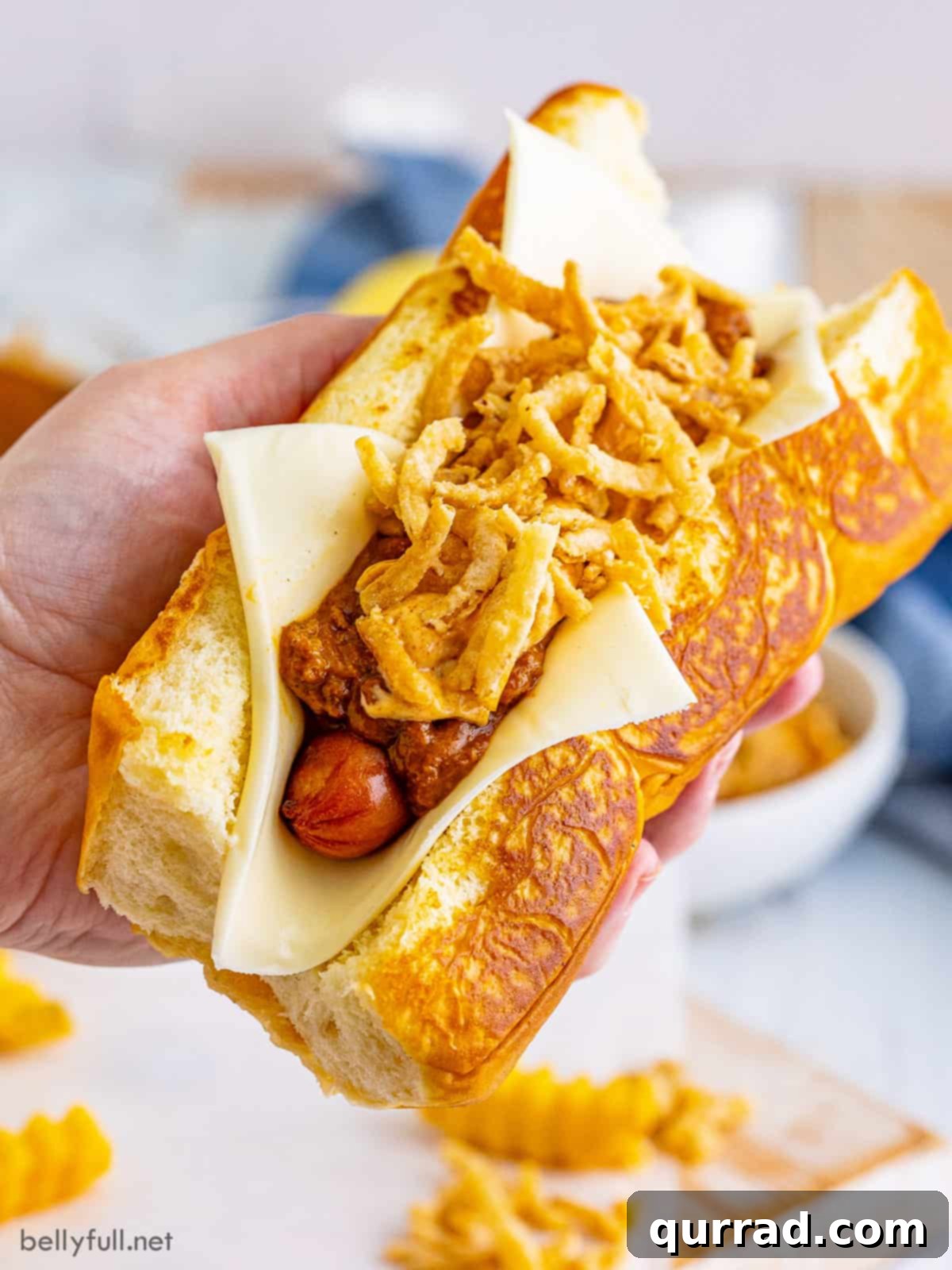 A hand holding a perfectly assembled gourmet chili dog with visible layers of chili, cheese, and aioli