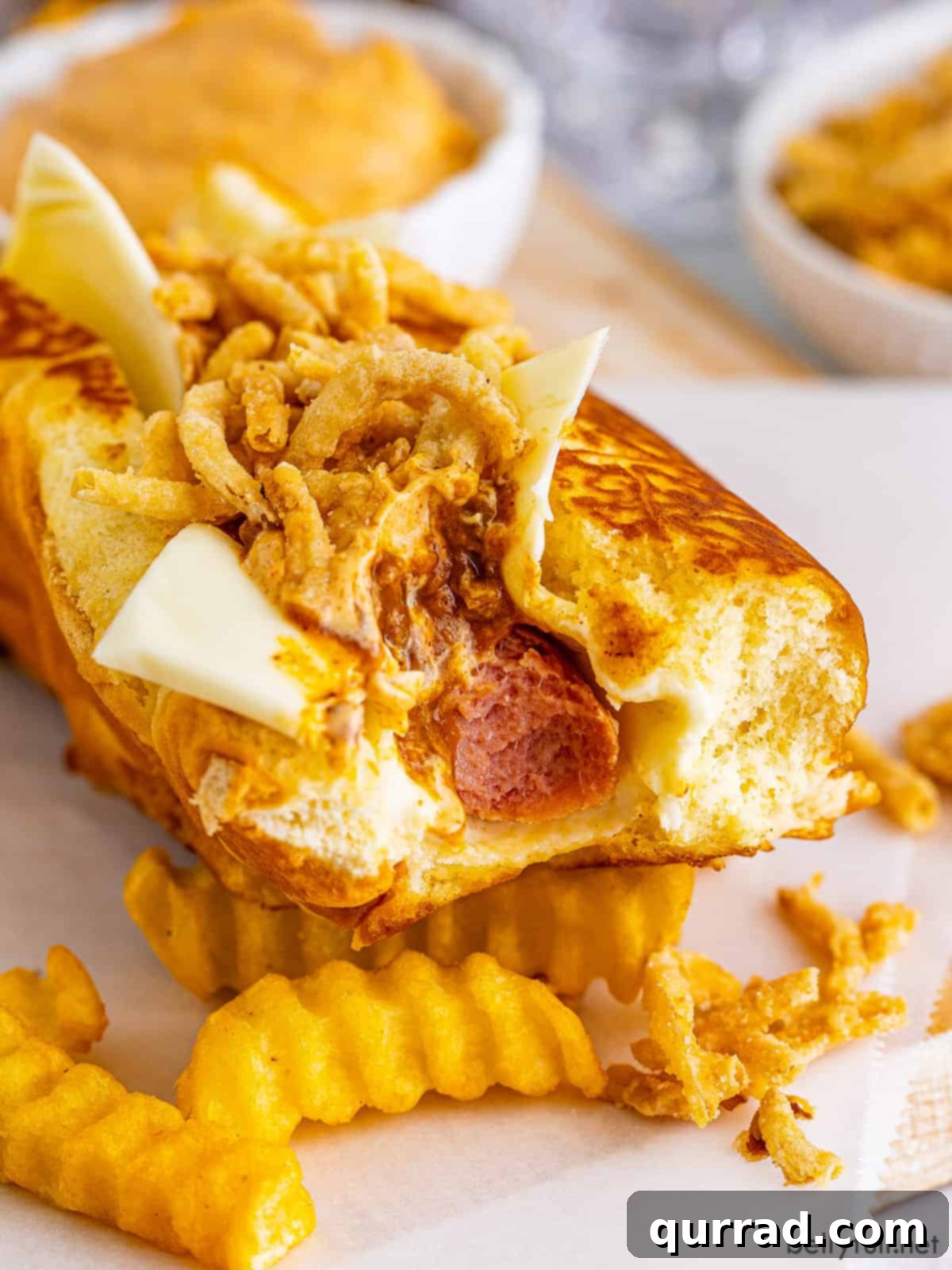A gourmet hot dog, topped with melted white American cheese, a rich chili, and crunchy fried onions, with a bite taken out