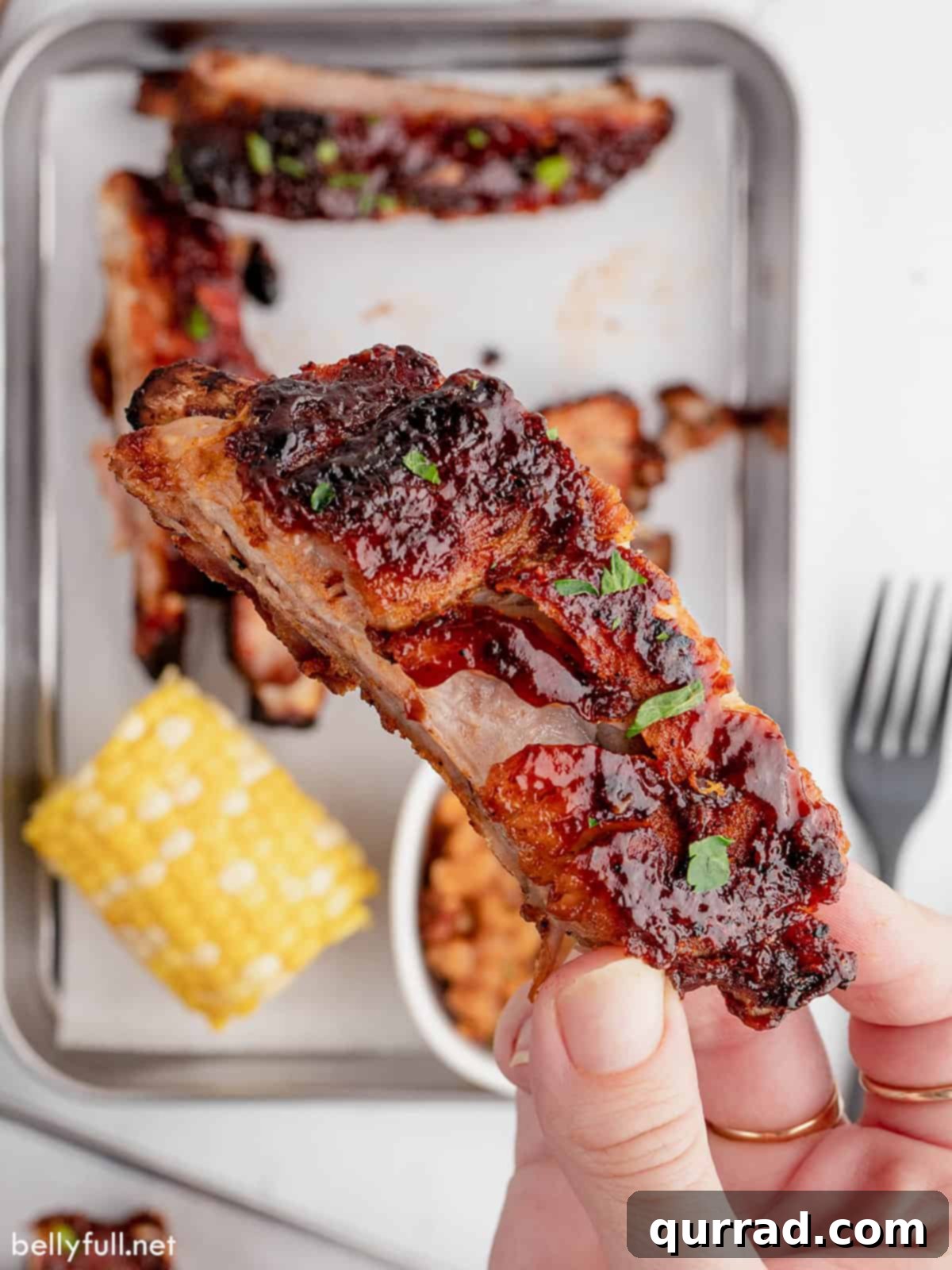 A hand holding a baby back rib over a baking sheet with other ribs, showing its tenderness