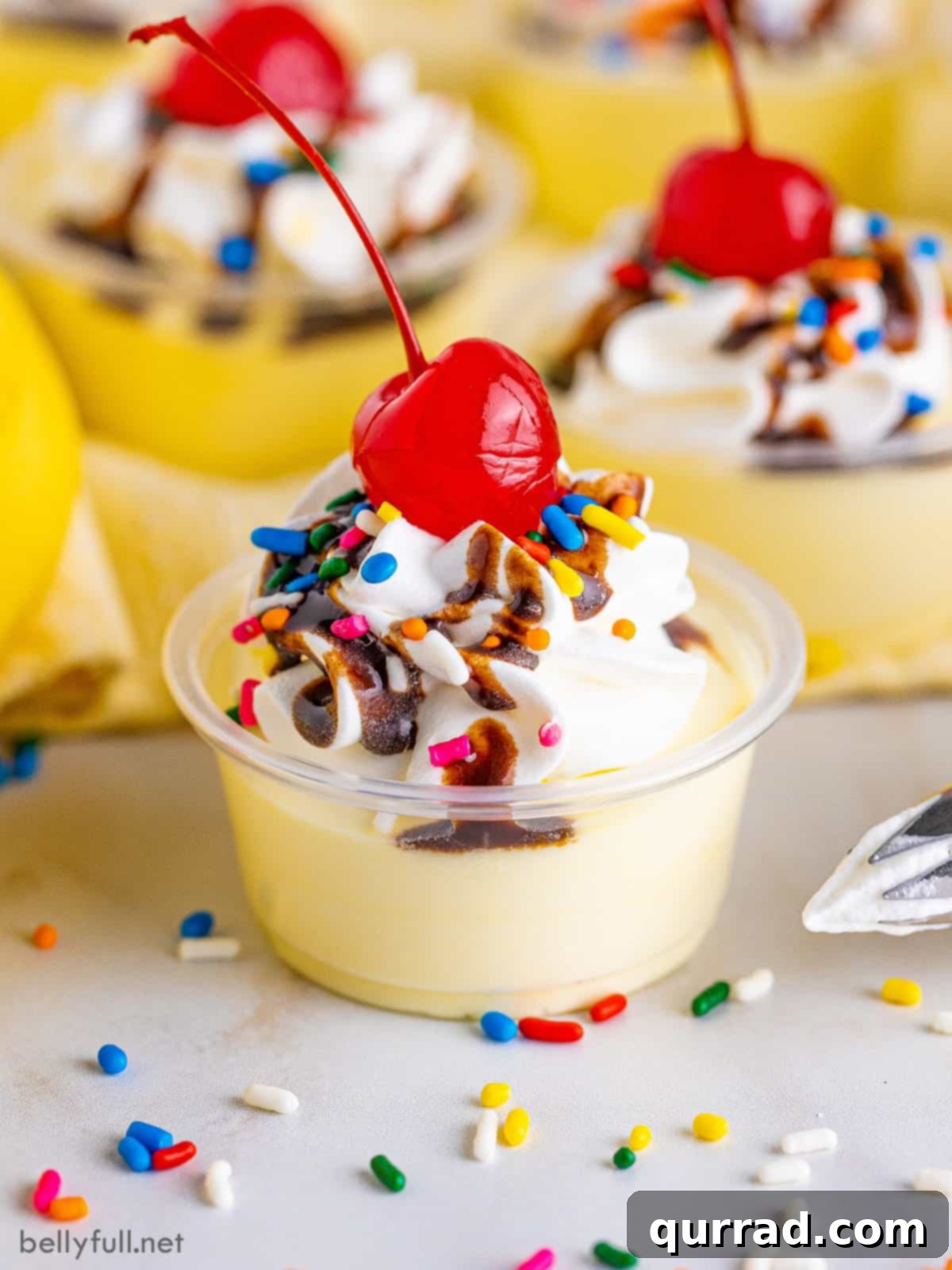Delicious Banana Split Pudding Shots ready for a party. A vibrant front view of several Banana Split Pudding Shots, each garnished with whipped cream, sprinkles, and a maraschino cherry.