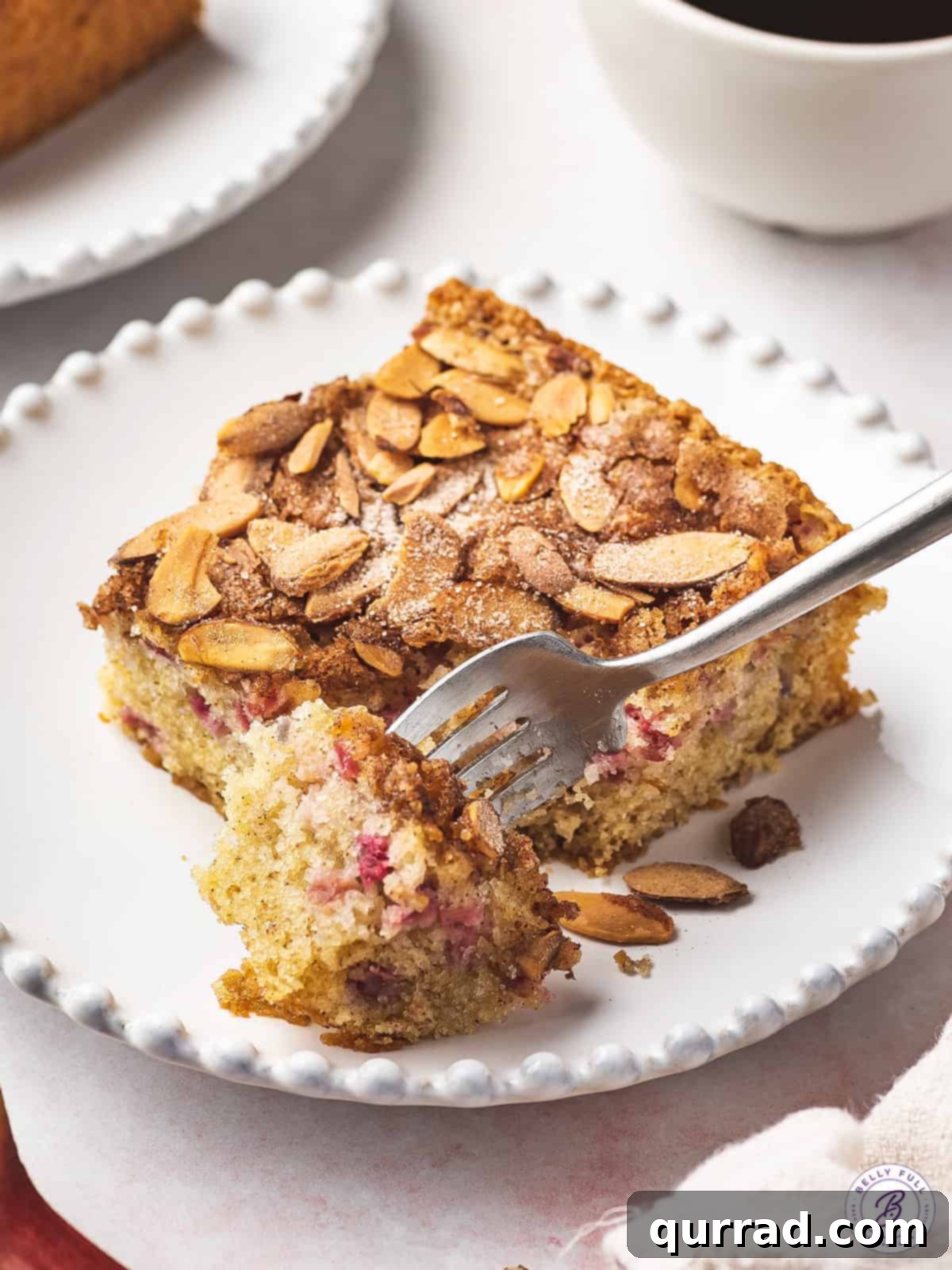 A tender slice of Rhubarb Coffee Cake with Almonds, showcasing its tempting crumb. A close-up shot of a slice of coffee cake on a white plate, with a fork cutting into it, highlighting the fluffy texture, moist interior, and visible rhubarb pieces.