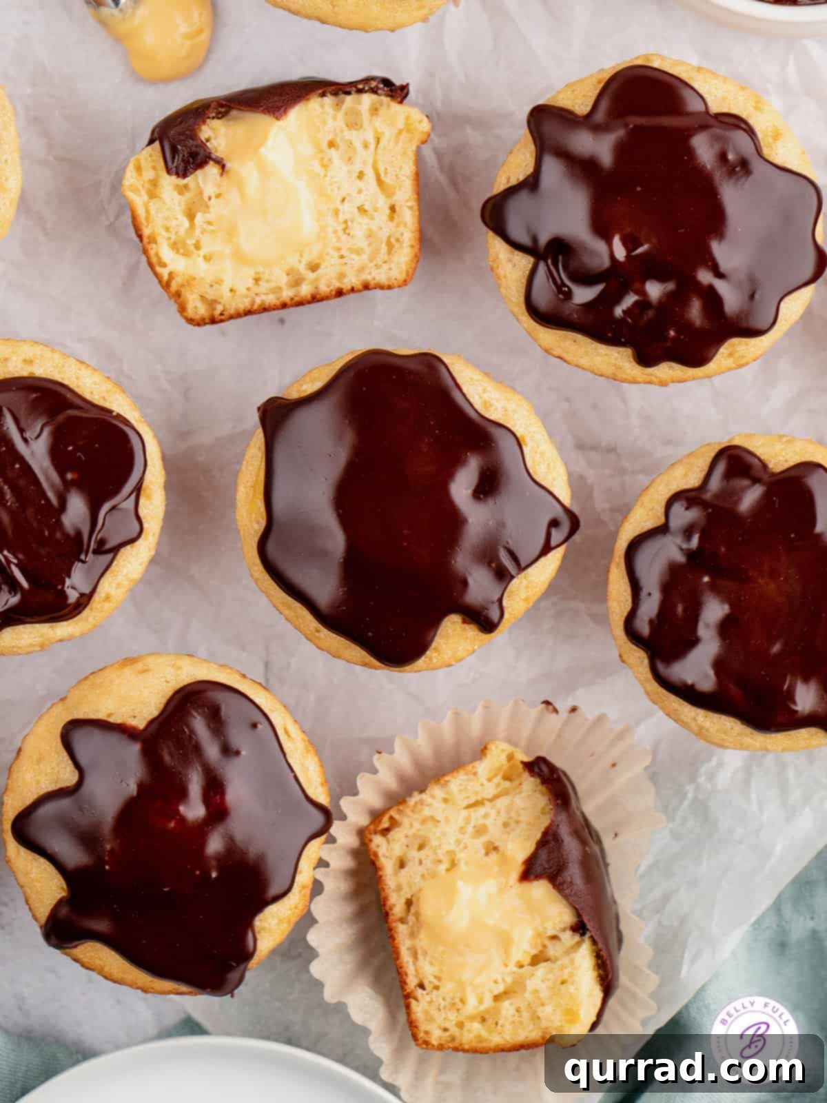 Overhead view of yellow cupcakes revealing a creamy vanilla filling and glossy chocolate ganache. An overhead shot of several Boston Cream Pie Cupcakes, some cut in half to reveal the creamy vanilla filling