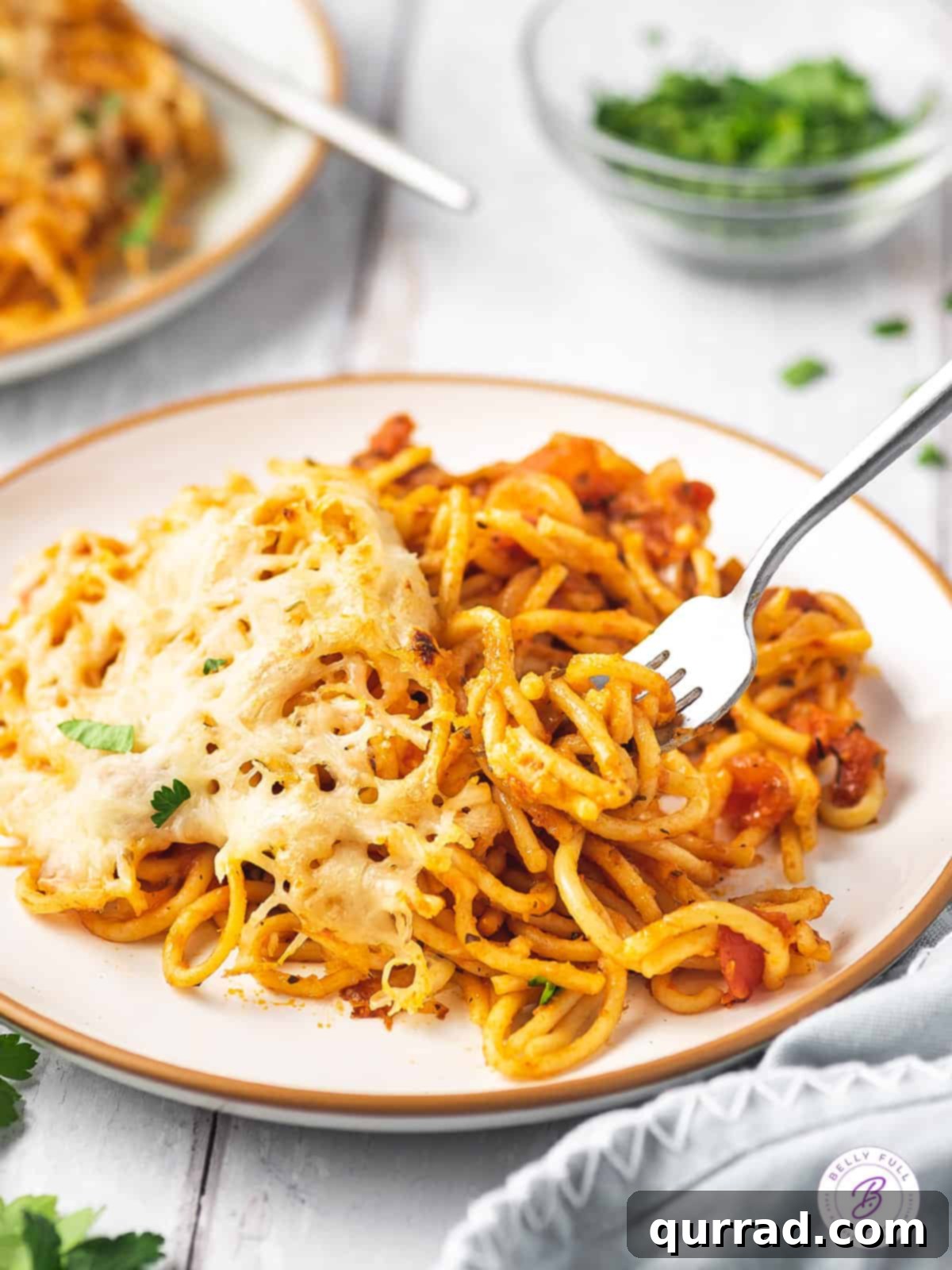 No pre-cooking needed! Just toss, add broth, and bake for amazing baked spaghetti. A portion of baked spaghetti on a white plate, served with a fork, showing melted cheese.