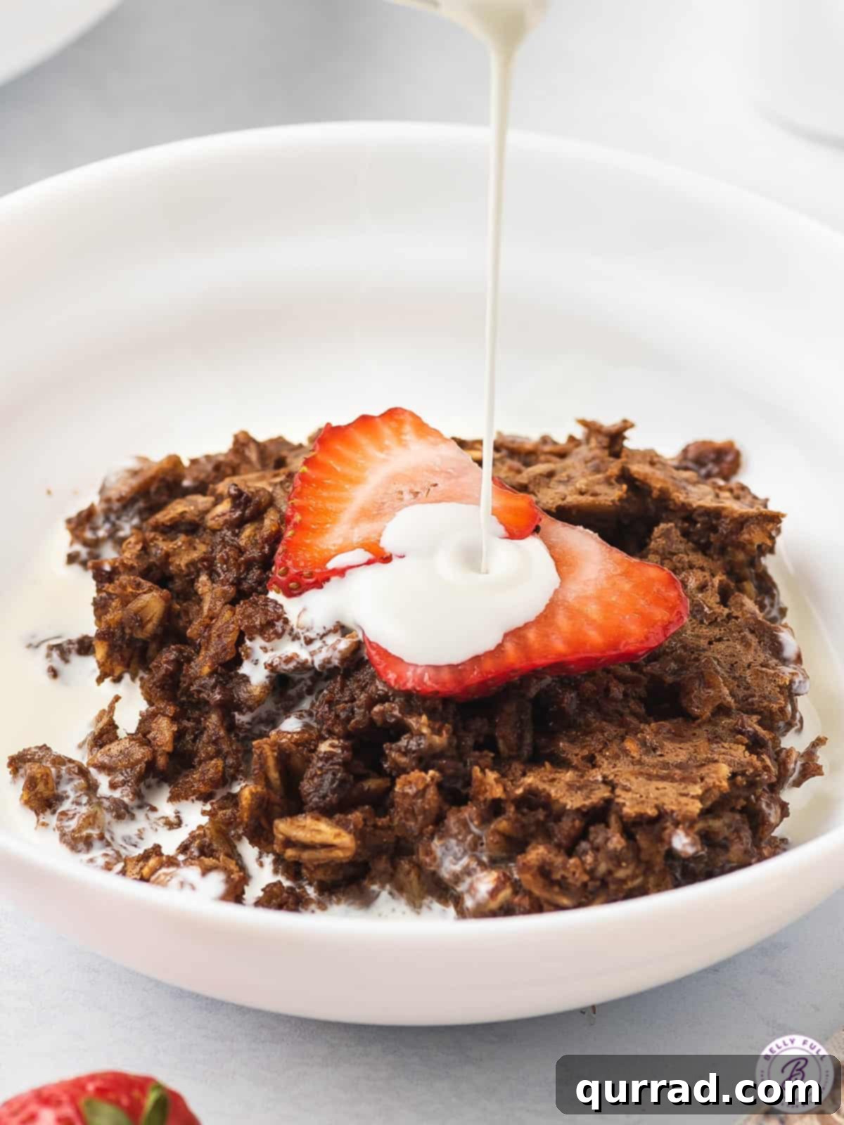 Heavy cream being elegantly drizzled over a warm serving of brownie baked oatmeal, garnished with fresh strawberries, highlighting the creamy topping.