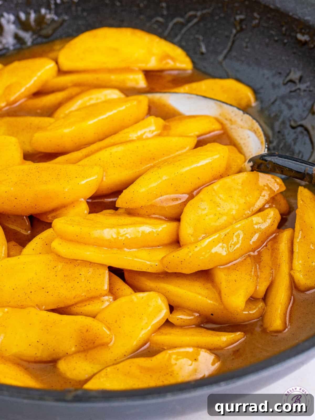 A large spatula gently stirring glistening fried apples in a stainless steel skillet, highlighting the caramelization and rich sauce.