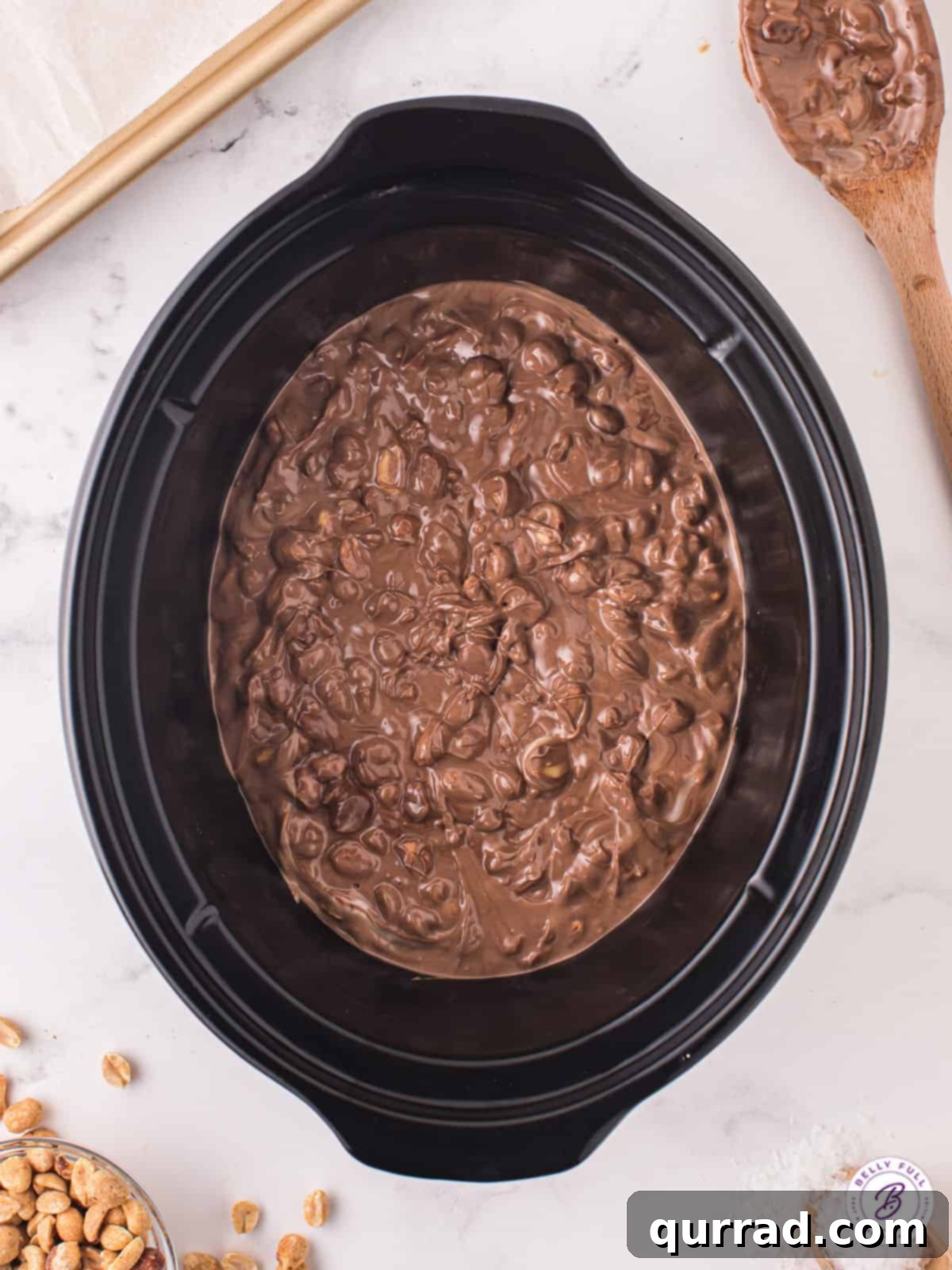 The slow cooking process, where chocolate and peanuts blend harmoniously. Melted chocolate and peanuts mixing gently inside a crockpot, creating the candy base.