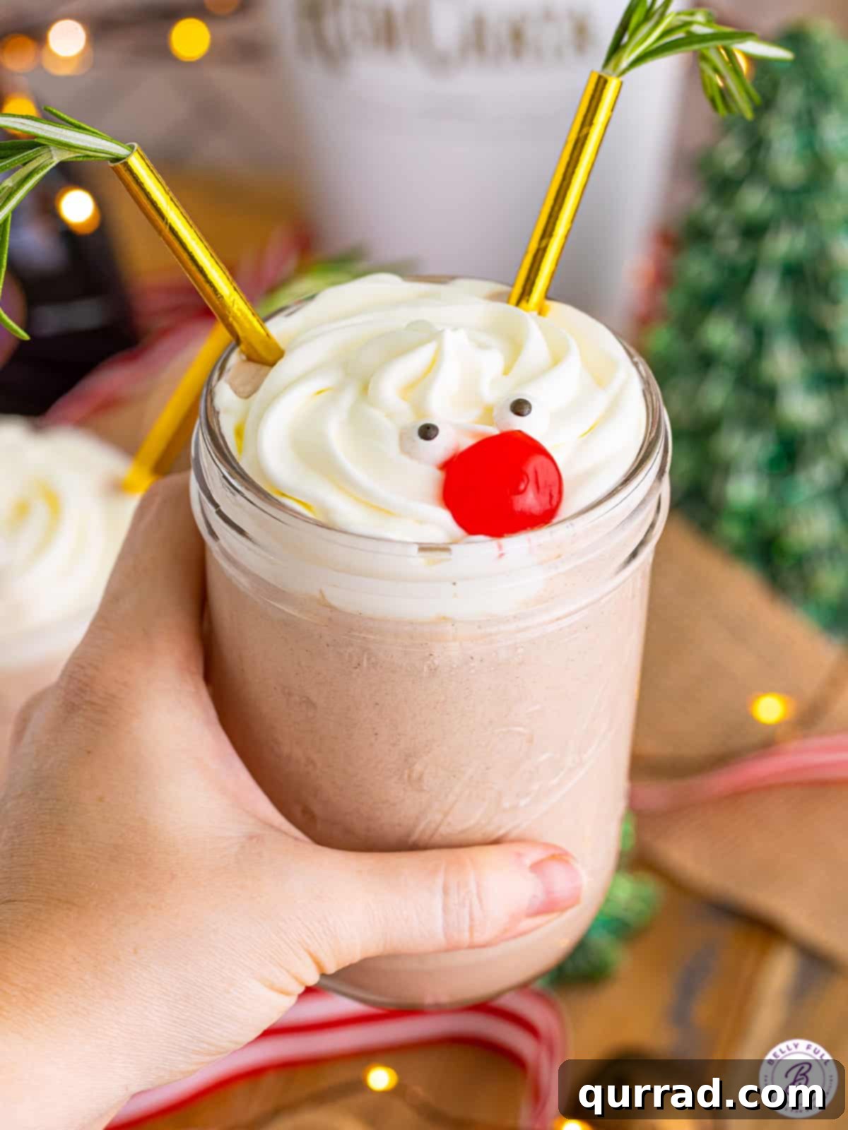 Made with 3 types of liquor and vanilla ice cream, this Drunk Rudolph Milkshake is the Christmas cocktail you didn't know you needed. A hand holding a garnished Drunk Rudolph Milkshake in a mason jar with a red cherry nose, candy eyes, and rosemary antlers.