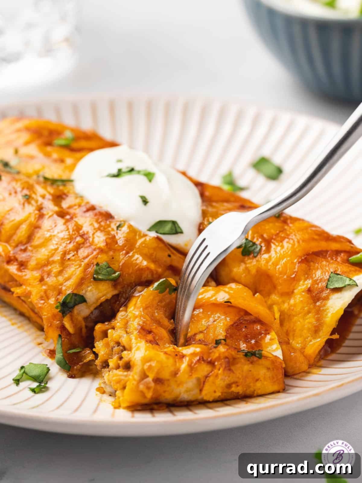 A fork cuts into a freshly baked ground beef enchilada on a white plate, revealing the rich filling and melted cheese.