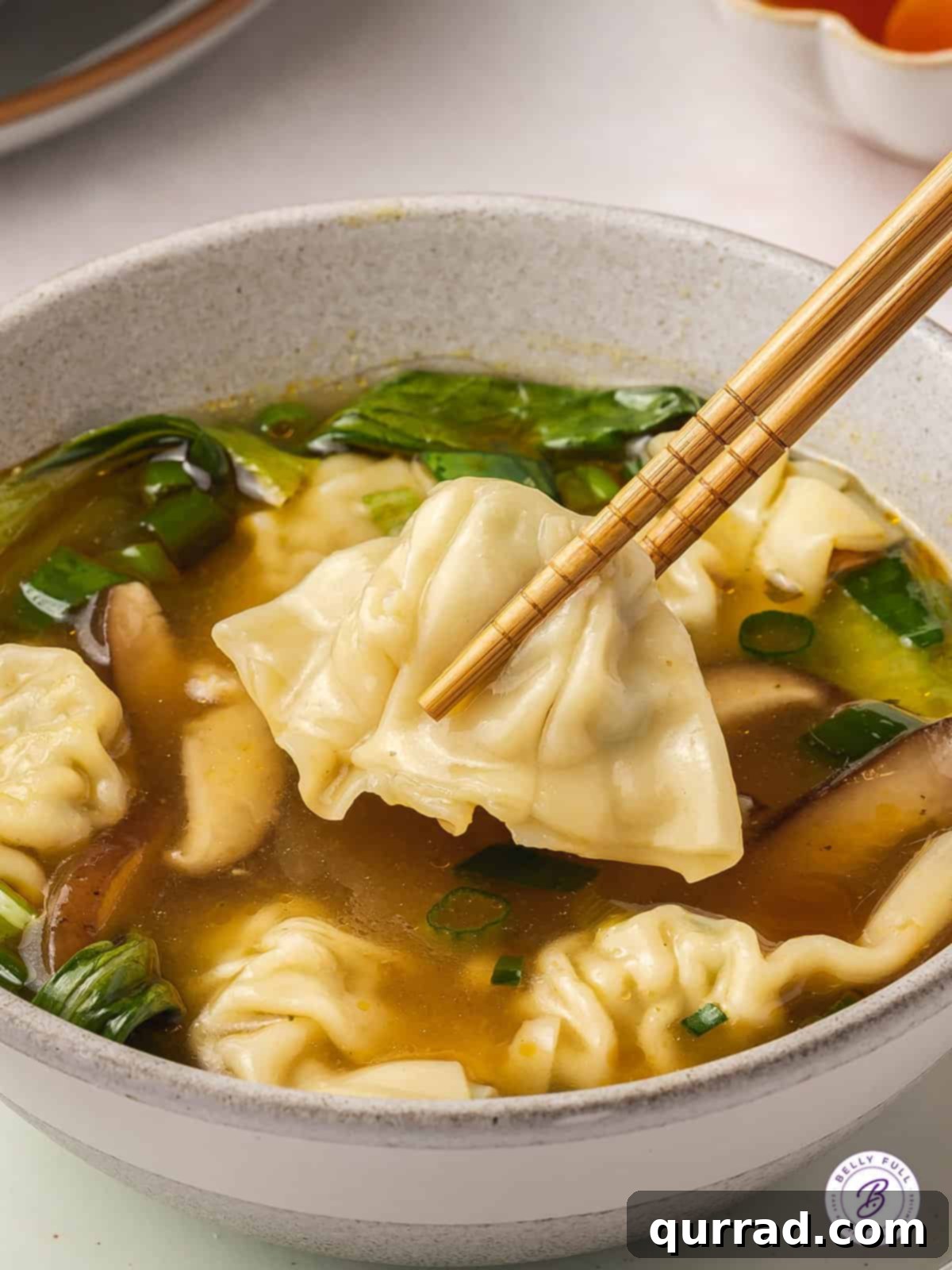 Delicious homemade Wonton Soup made with frozen wontons, bok choy, and mushrooms, ready in under 30 minutes. A single mini wonton held by chopsticks above a bowl of homemade wonton soup, emphasizing the delicious filling.