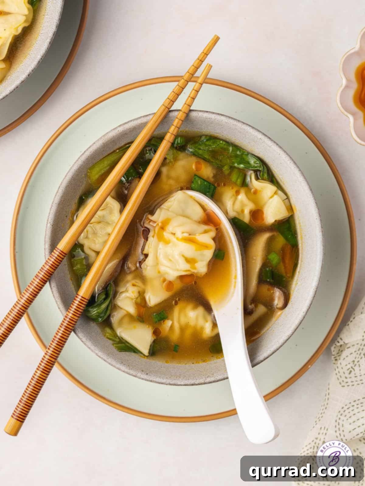 Cloud Dumplings in Golden Broth 6 Overhead view of a beautifully garnished bowl of homemade wonton soup with a white spoon and chopsticks.