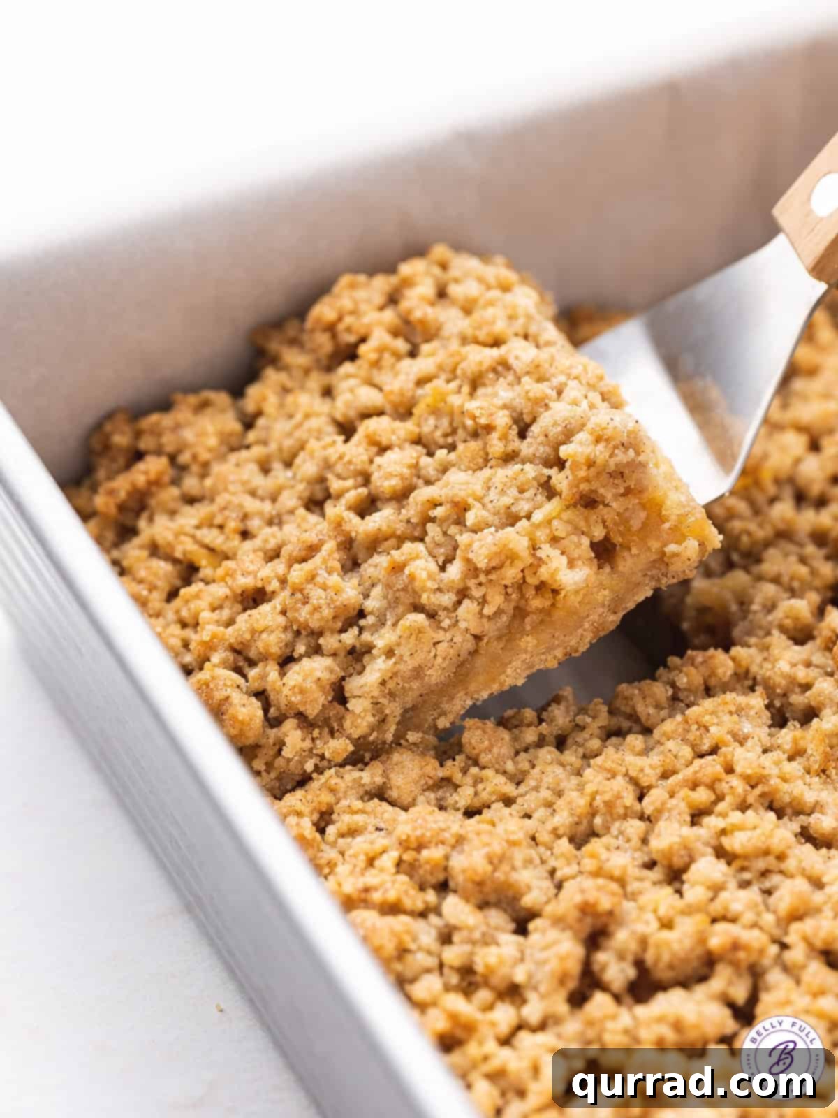 Apple Crumble Bars 9 A warm, golden apple pie bar being gently lifted from a baking pan with a spatula, showcasing its thick apple filling and crisp crumble topping, ready to be served.