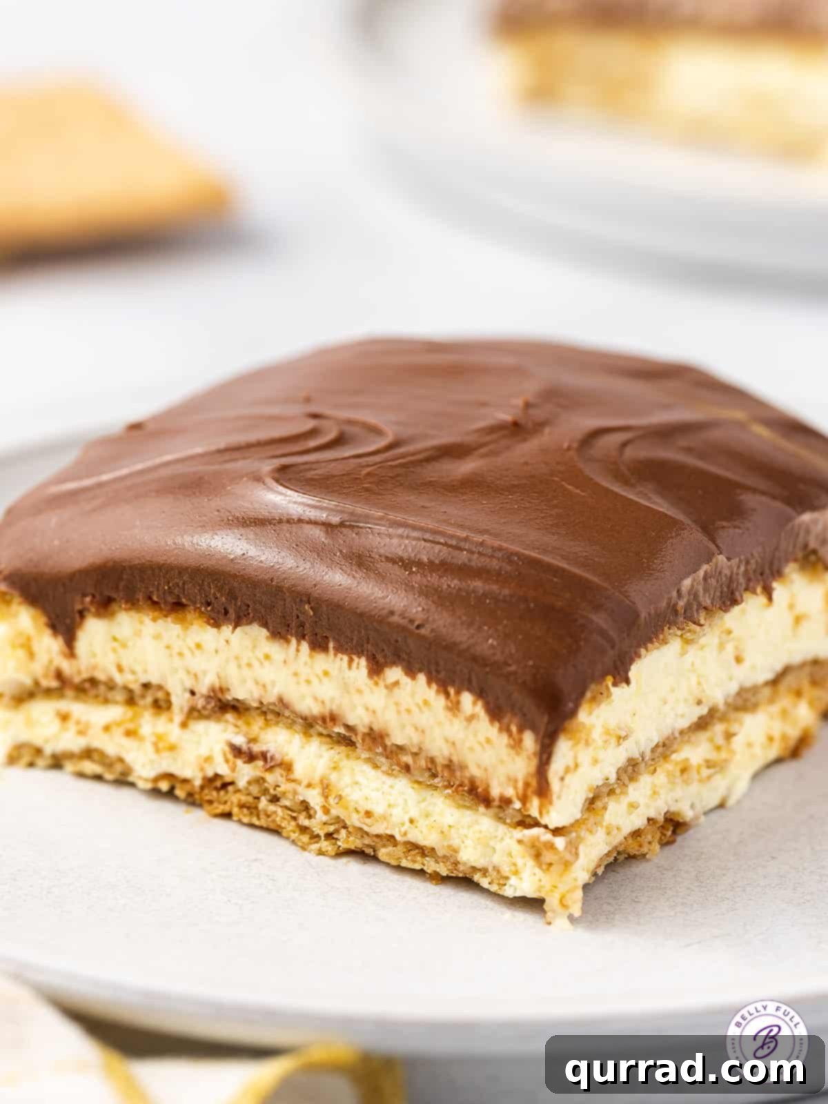 This Chocolate Eclair Dessert tastes like the best combination of Boston cream pie and a chocolate eclair. Just 5 ingredients! A perfectly portioned slice of No-Bake Chocolate Eclair Cake on a white plate, showcasing its distinct layers of graham cracker, creamy vanilla filling, and rich chocolate topping.