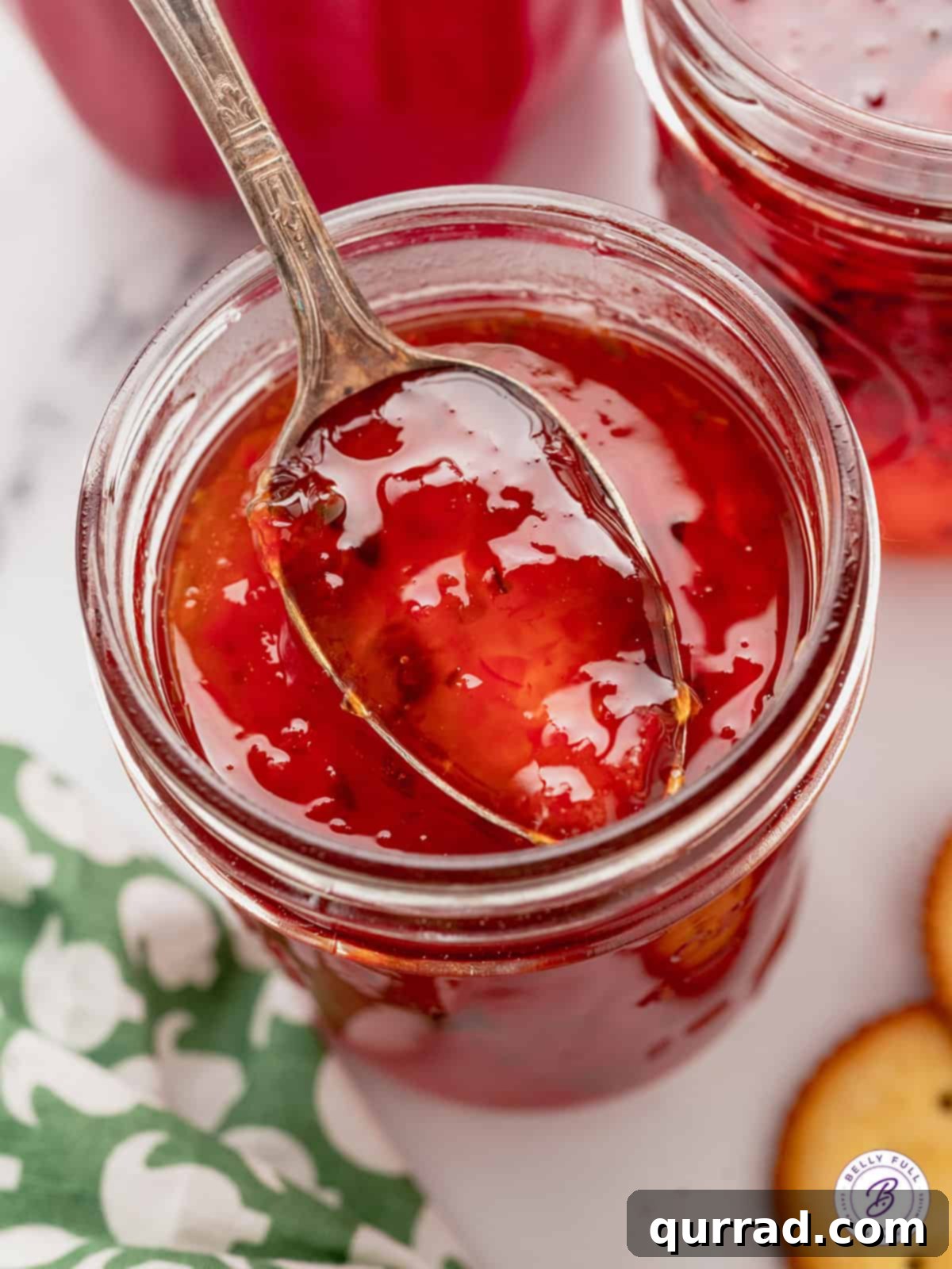 A spoon in a jar of red pepper jelly, showcasing its vibrant color and smooth texture.