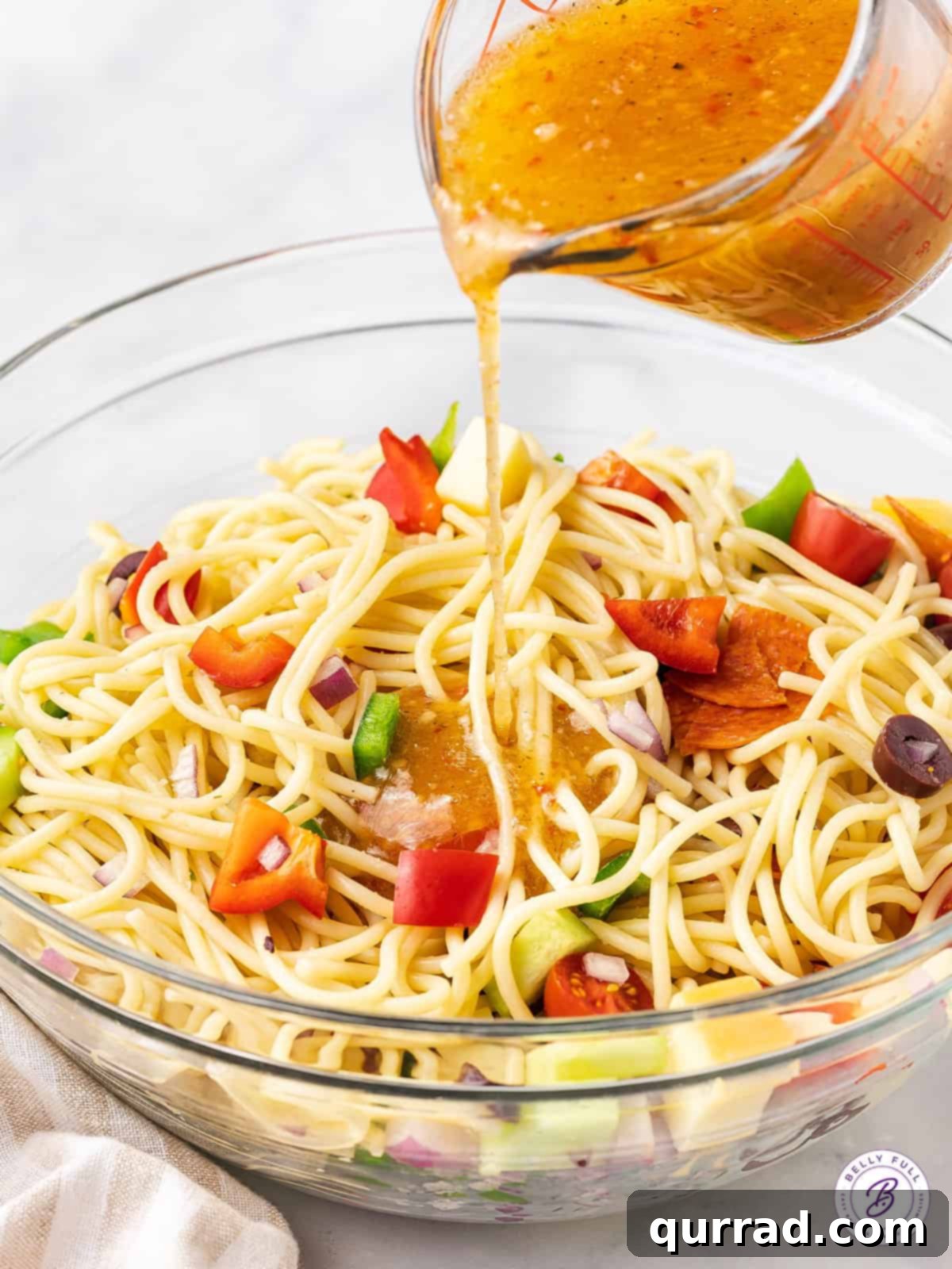 Italian dressing coats spaghetti salad: The secret to a zesty, flavorful pasta dish. A hand pouring zesty Italian dressing over a vibrant bowl of spaghetti pasta salad ingredients, ensuring even coating.
