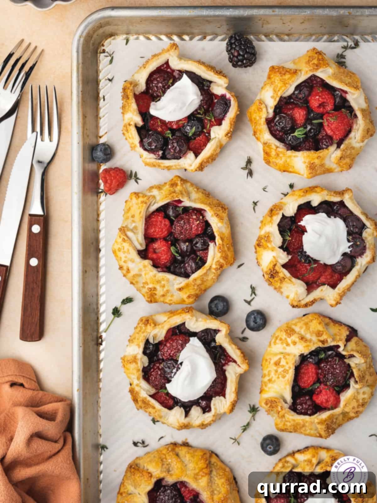 Made with strawberries, blueberries, raspberries, and black berries in a pie crust, these Mini Mixed Berry Galettes are elegant & easy. Overhead view of eight golden-brown mini galettes arranged neatly on a parchment-lined baking sheet, ready to be served.
